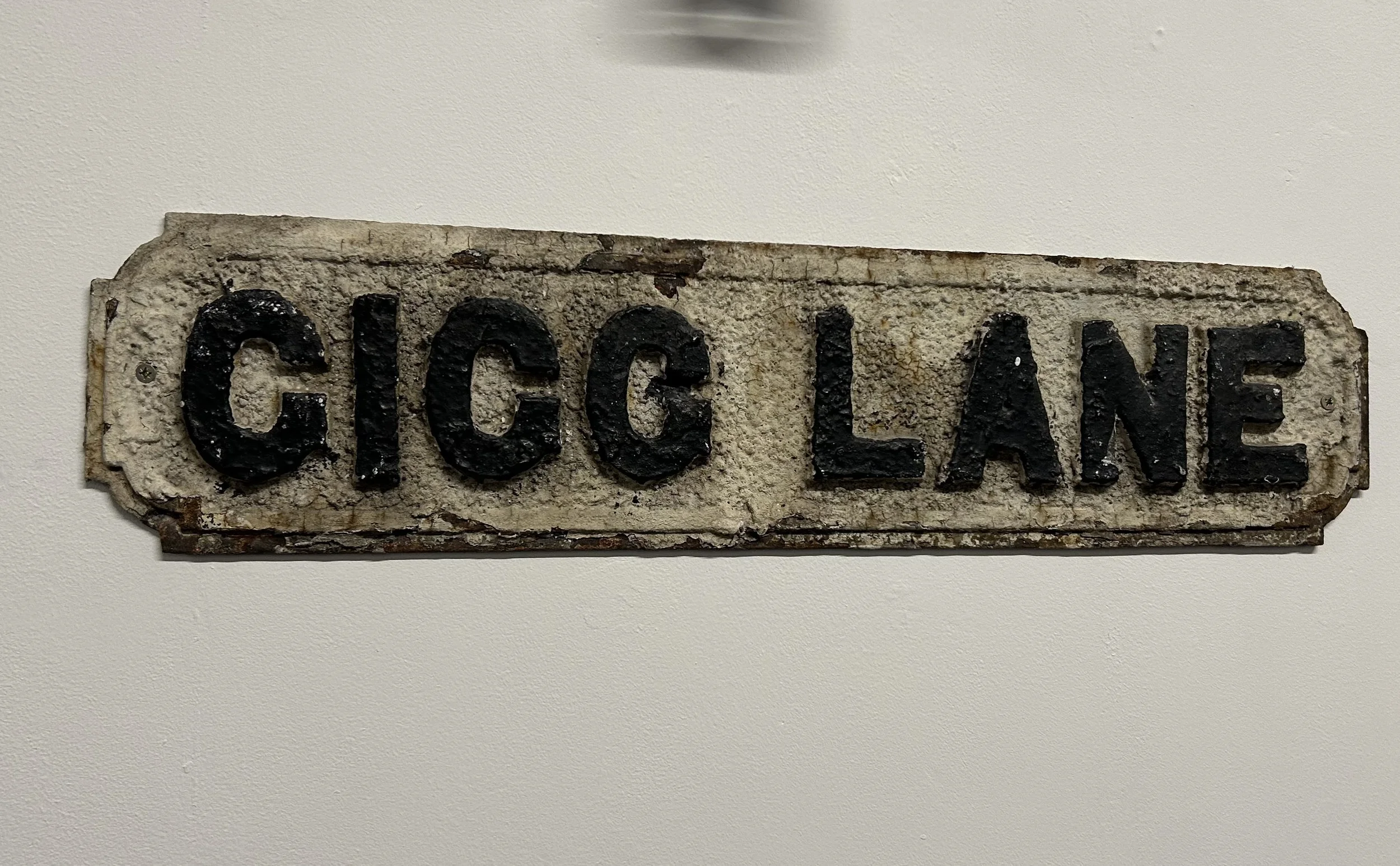 A weathered, vintage street sign reading 'GICG LANE' painted in black on a white, rusty metal background.