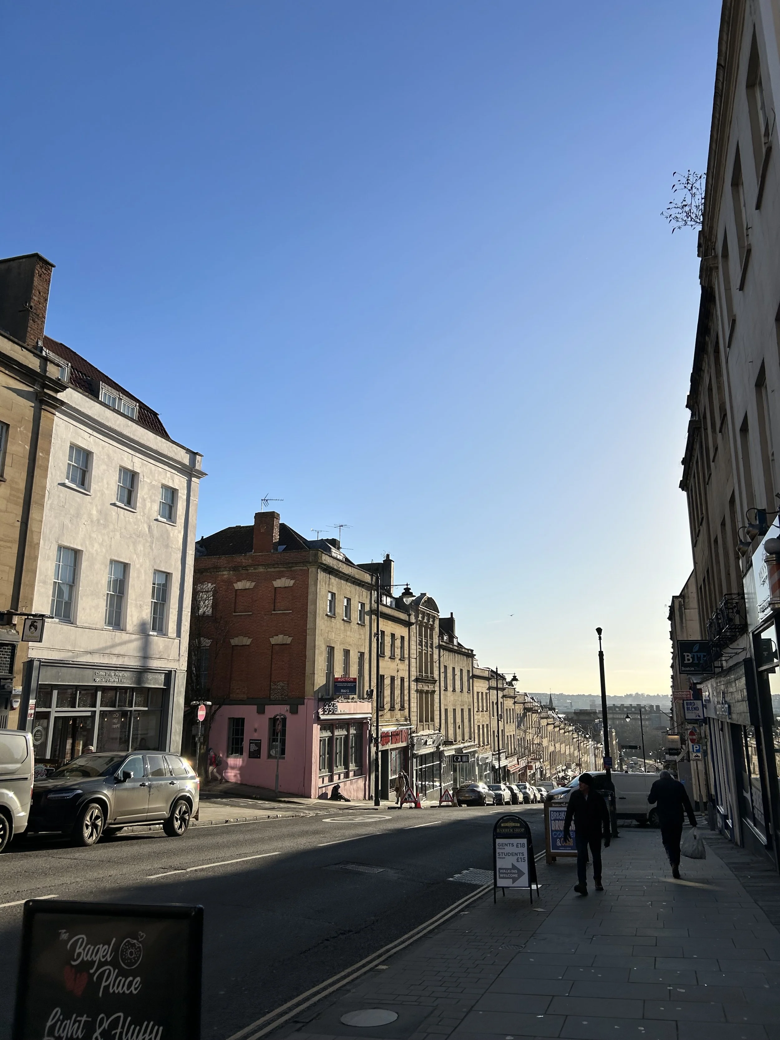 A city street scene with historic buildings, parked cars, two pedestrians walking on the sidewalk, and a clear blue sky overhead.