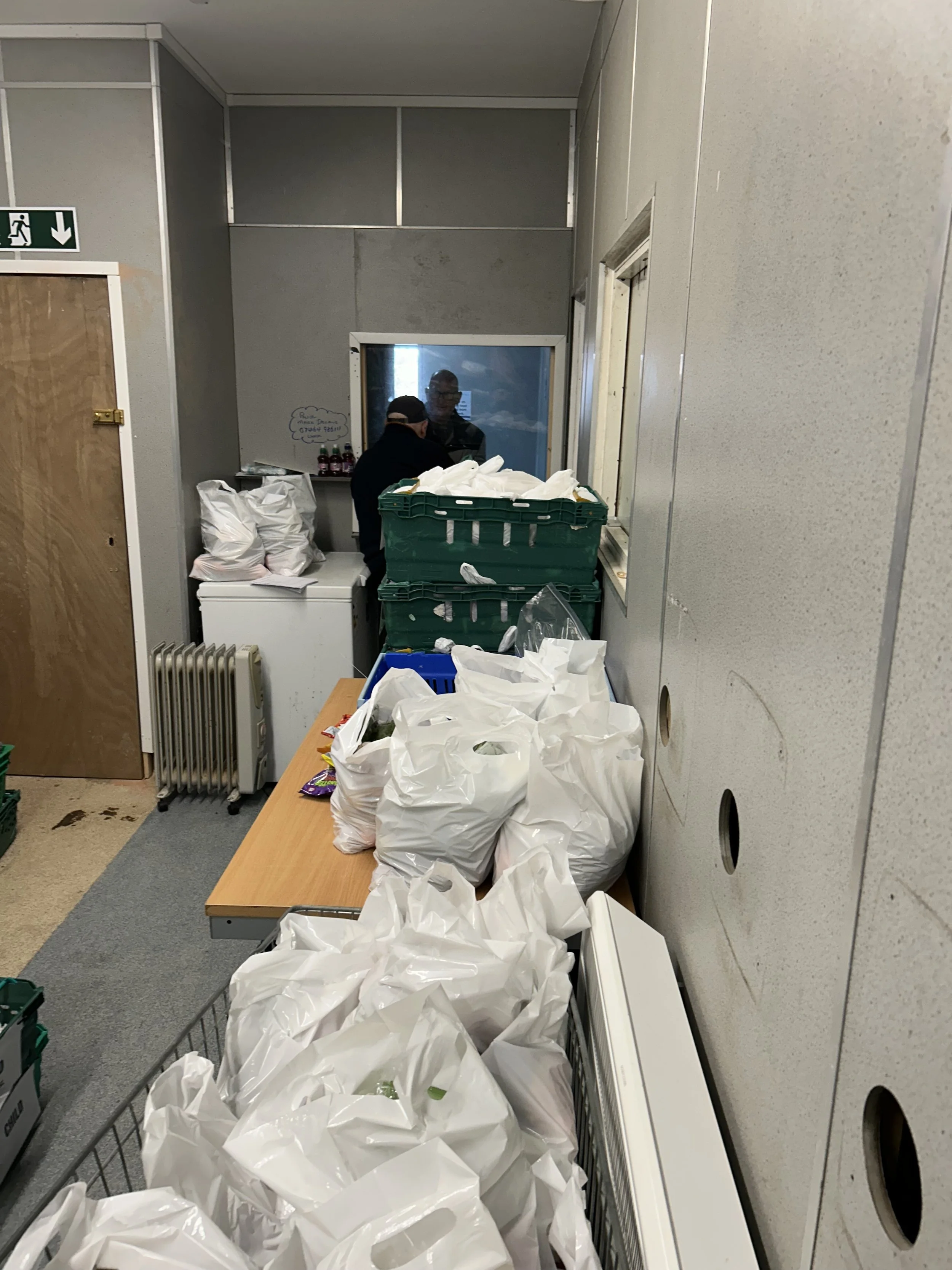 Room with a table or counter filled with white plastic grocery bags and stacked green crates, two men in dark clothing near a window or opening, and various objects including bottles and a whiteboard with writing.