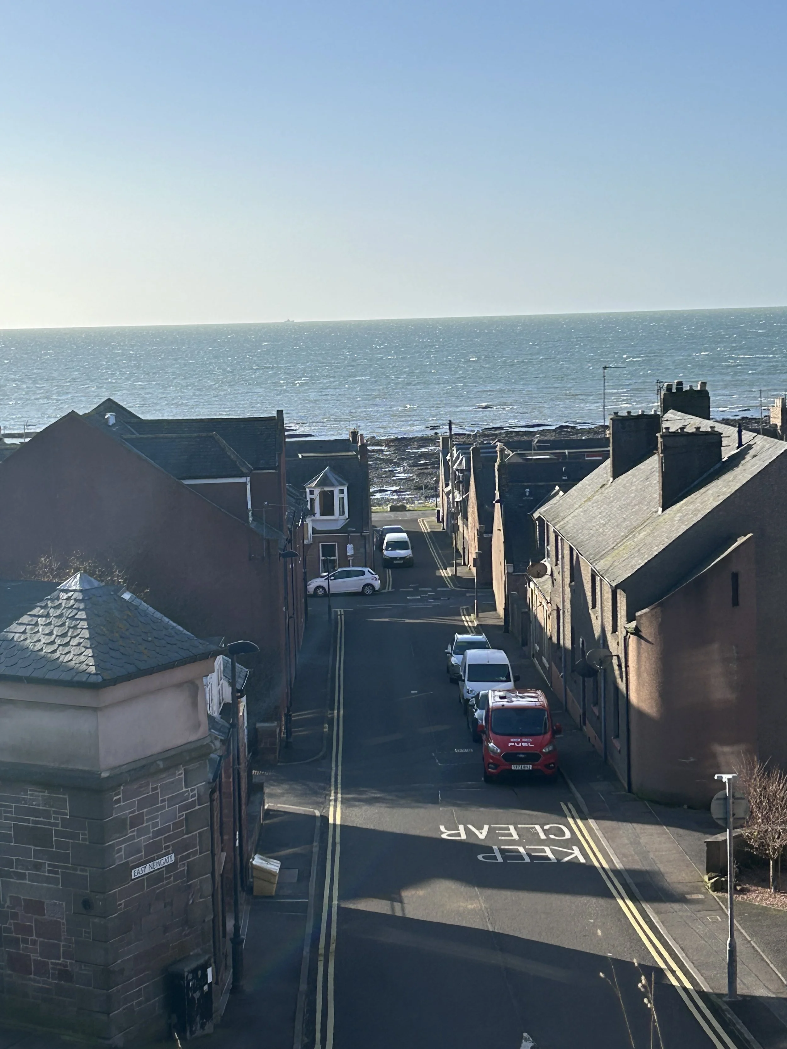 A narrow street leading to the ocean, with cars parked along the sides and houses on either side. The street has markings that say 'KEEP CLEAR.' The ocean and sky are visible in the background.