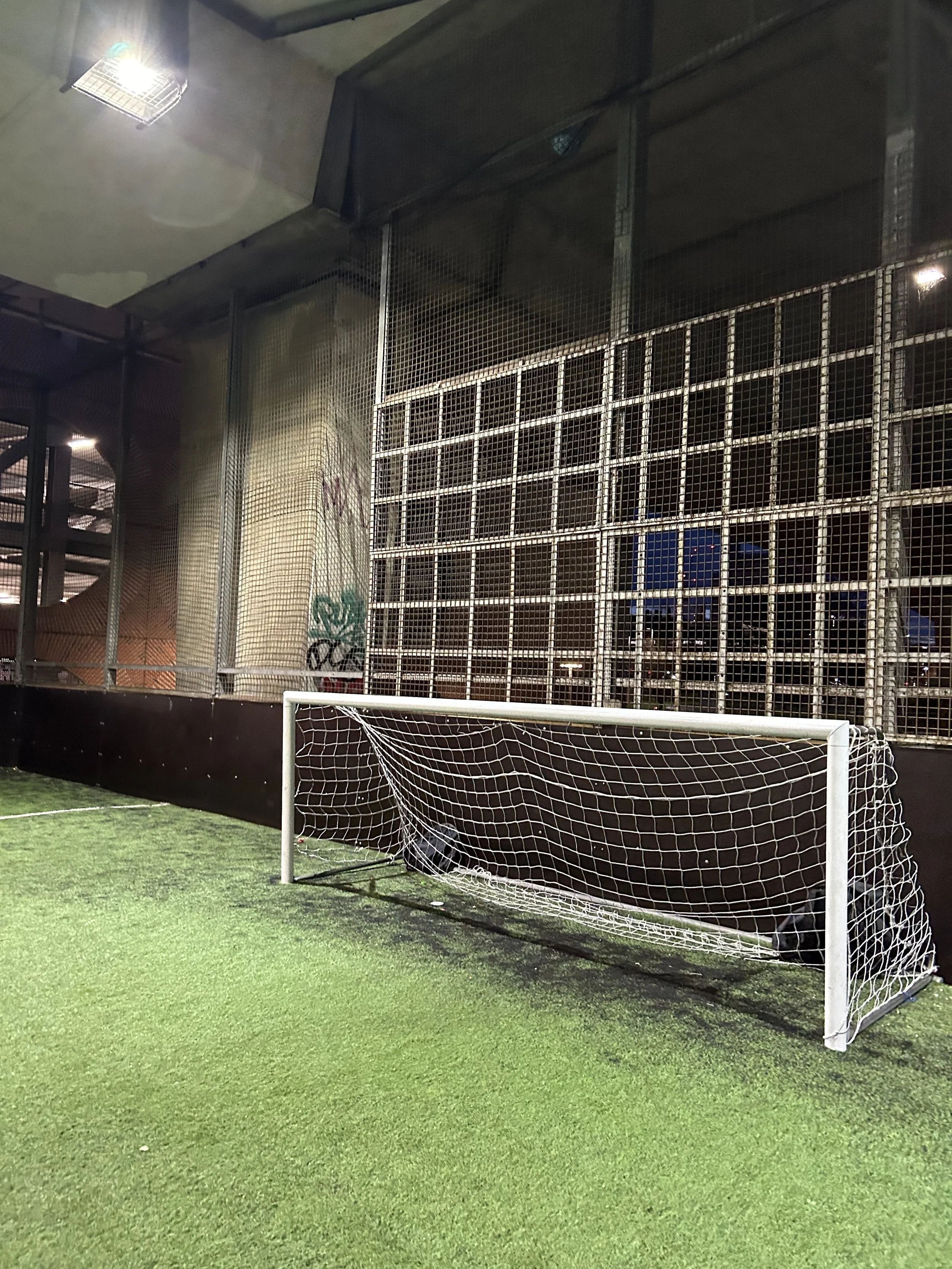 Indoor soccer goal on artificial turf with graffiti-covered wall and metal grid structure in background.