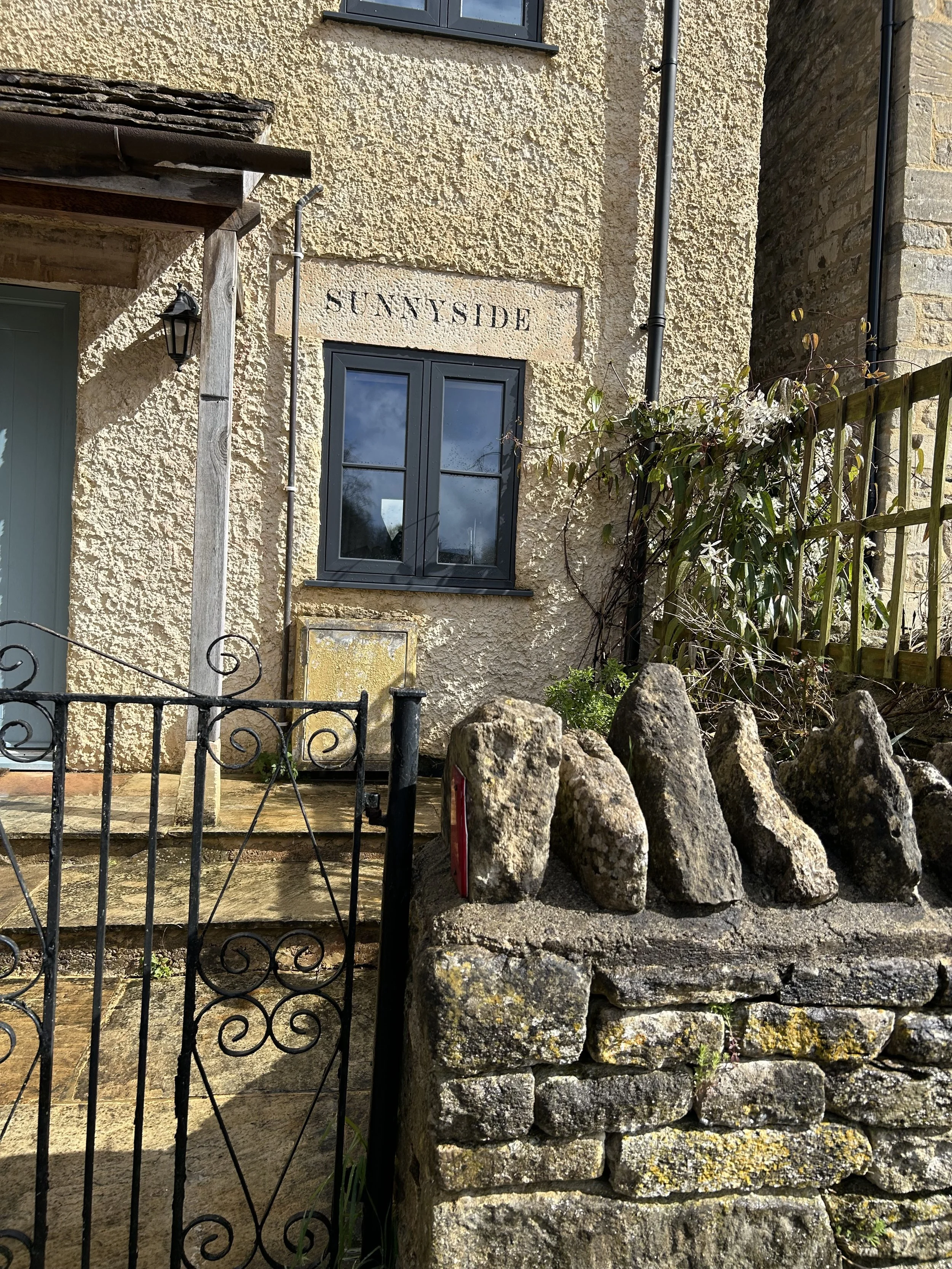 A stone house with a sign reading "Sunnyside" above a black-framed window. There is a small stone wall with rocks on top, a black iron gate, and a wooden fence on the right side. A light fixture is attached to the house, and there are some plants gro