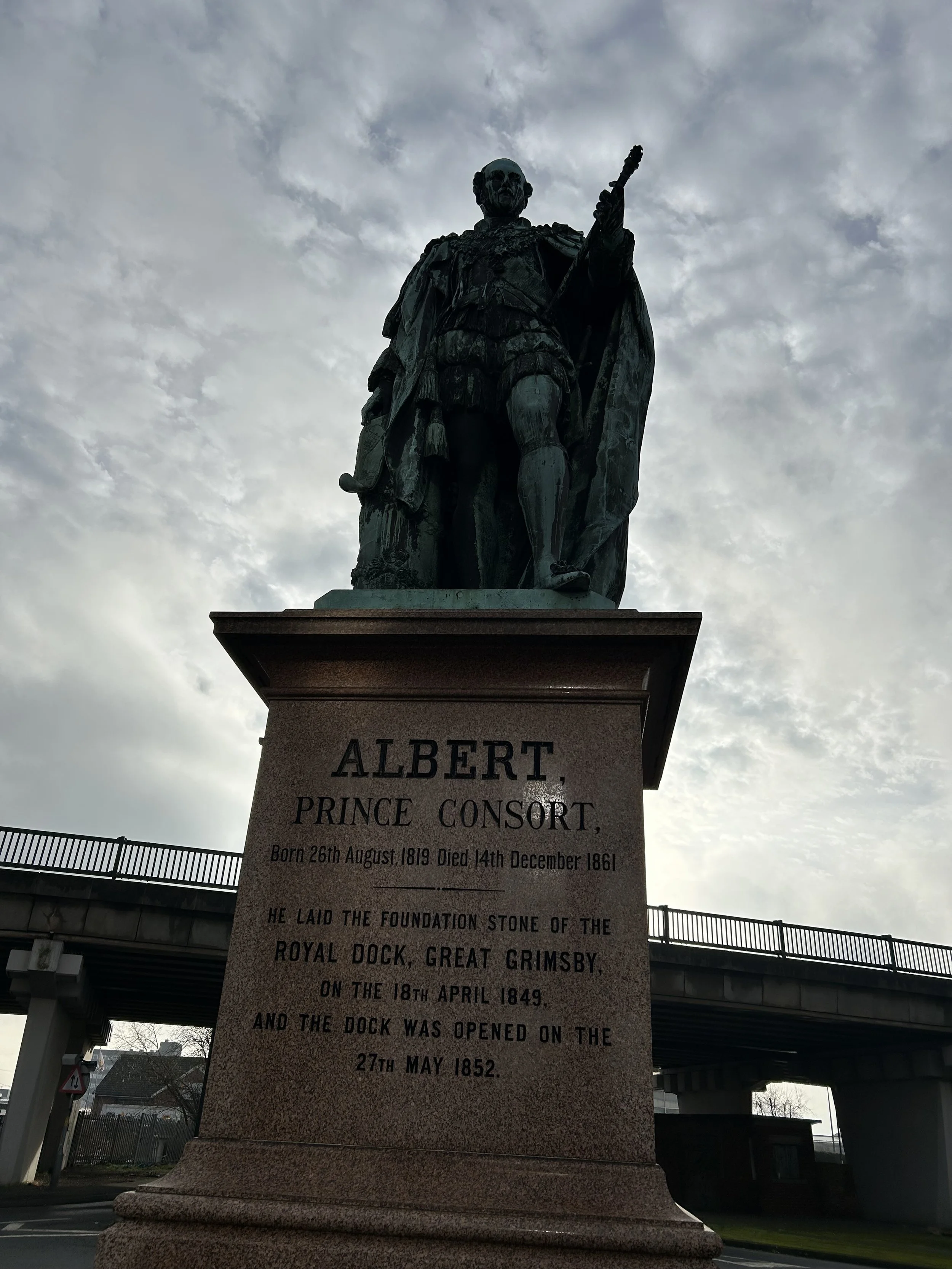 Statue of Albert, Prince Consort, on a stone pedestal with engraved inscription, located under a cloudy sky near a bridge.
