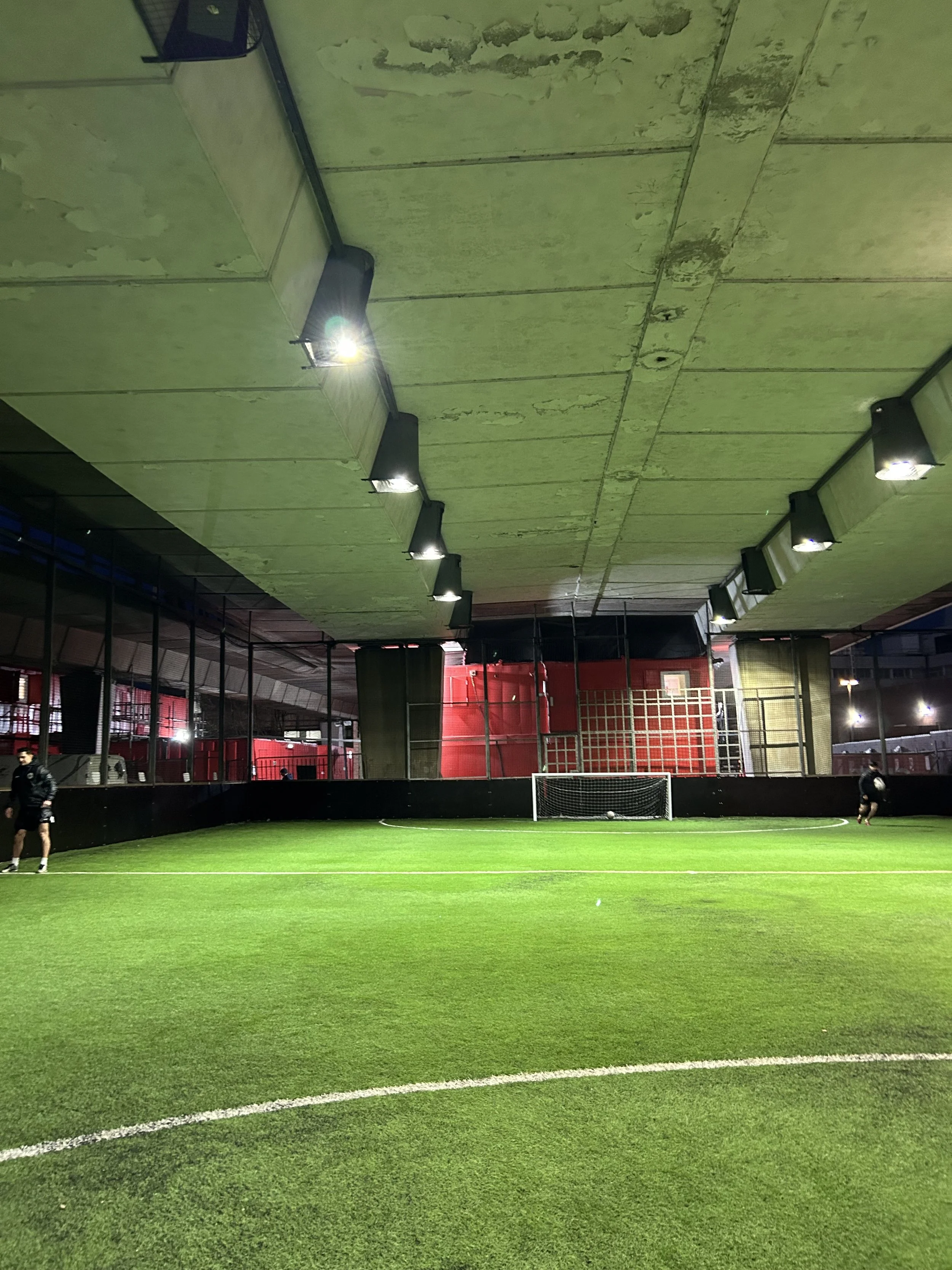Indoor soccer field with players practicing, artificial turf, goalpost, and ceiling lights.