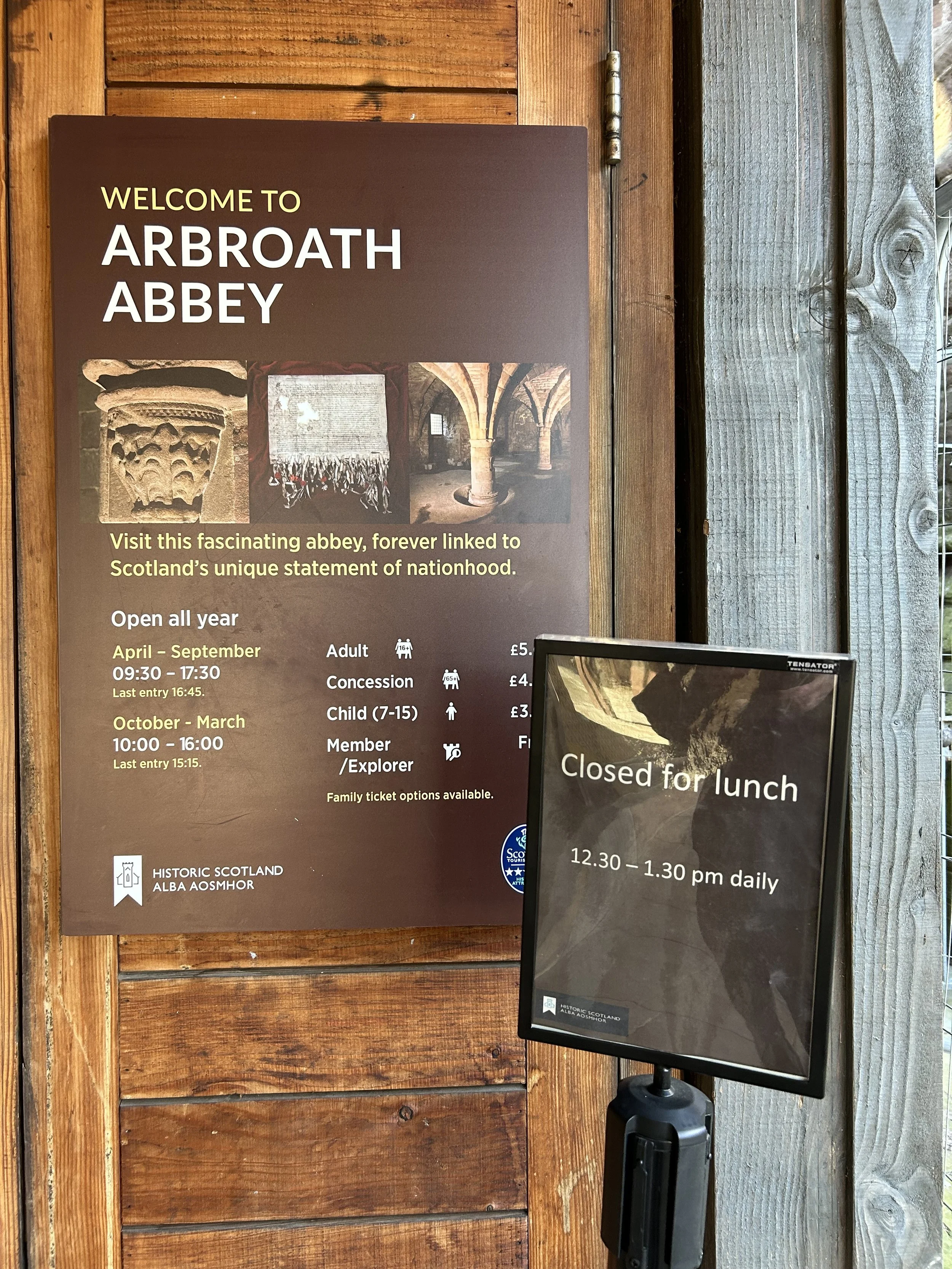 Signboard at Arbroath Abbey with details about visiting hours, ticket prices, and closure for lunch from 12:30 to 1:30 pm daily.