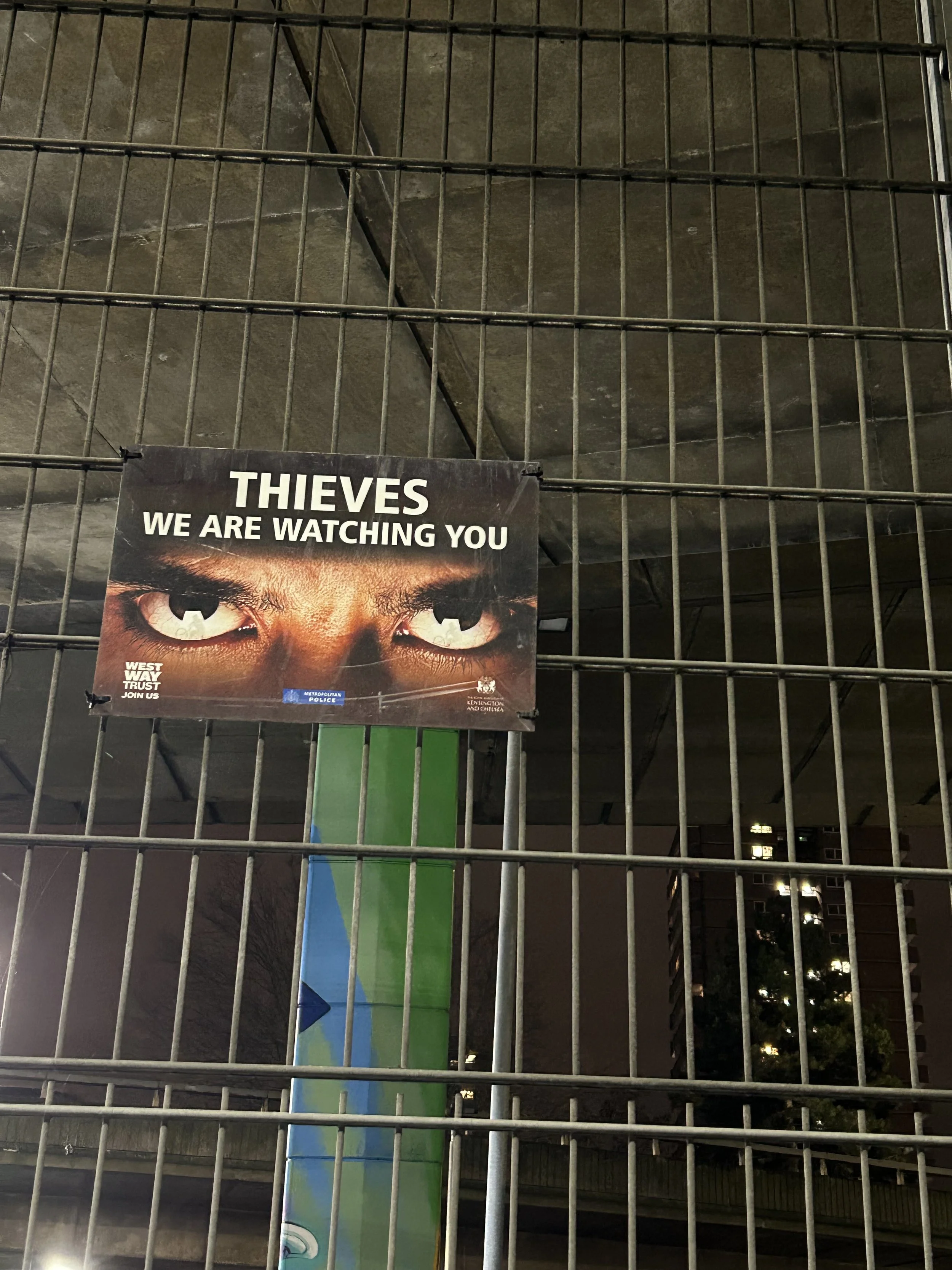 A metal fence with a surveillance warning sign showing intense eyes and the message 'Thieves, we are watching you,' at night in an urban setting.