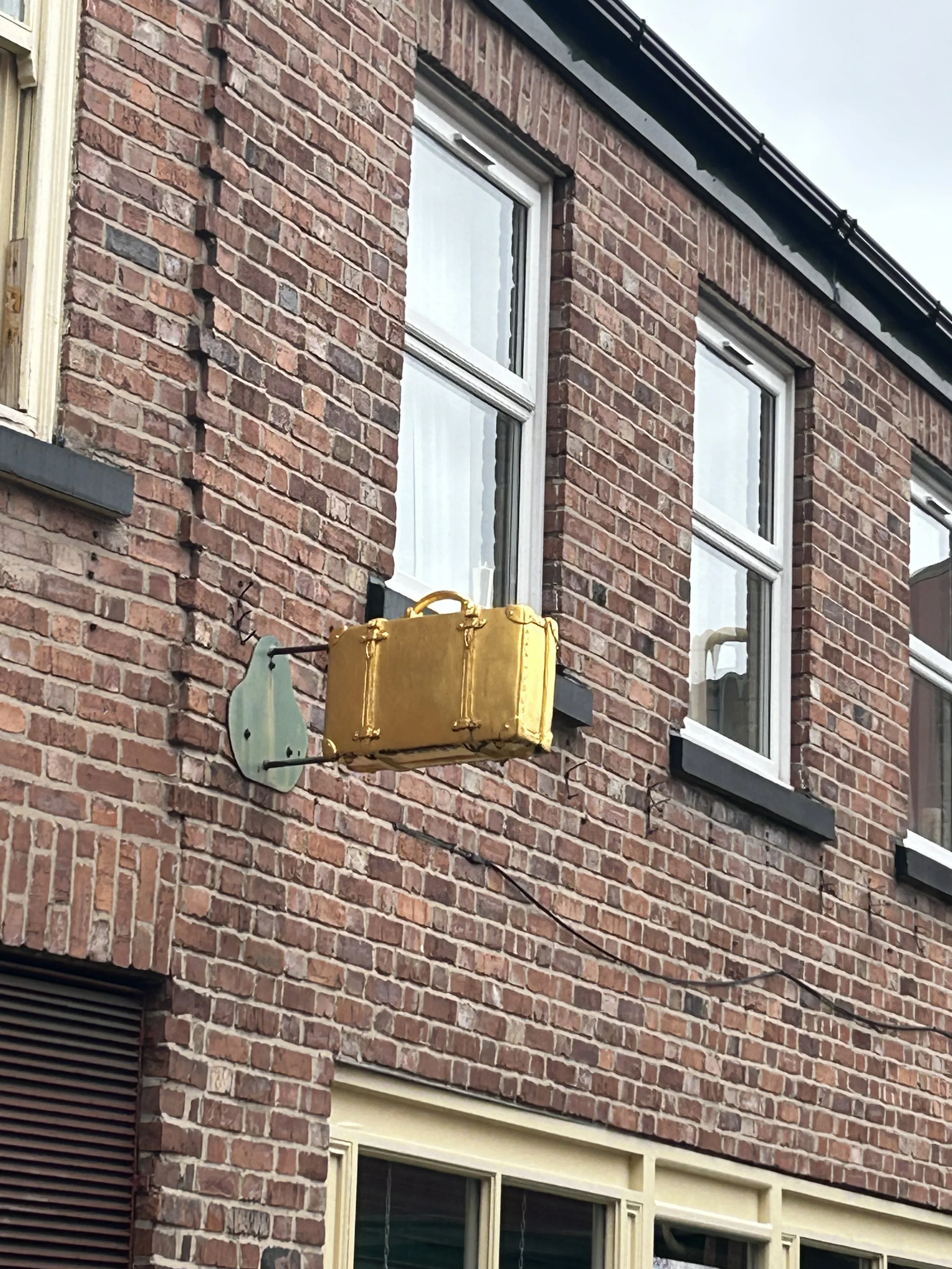 A vintage yellow suitcase hanging outside a brick building with three window panes.