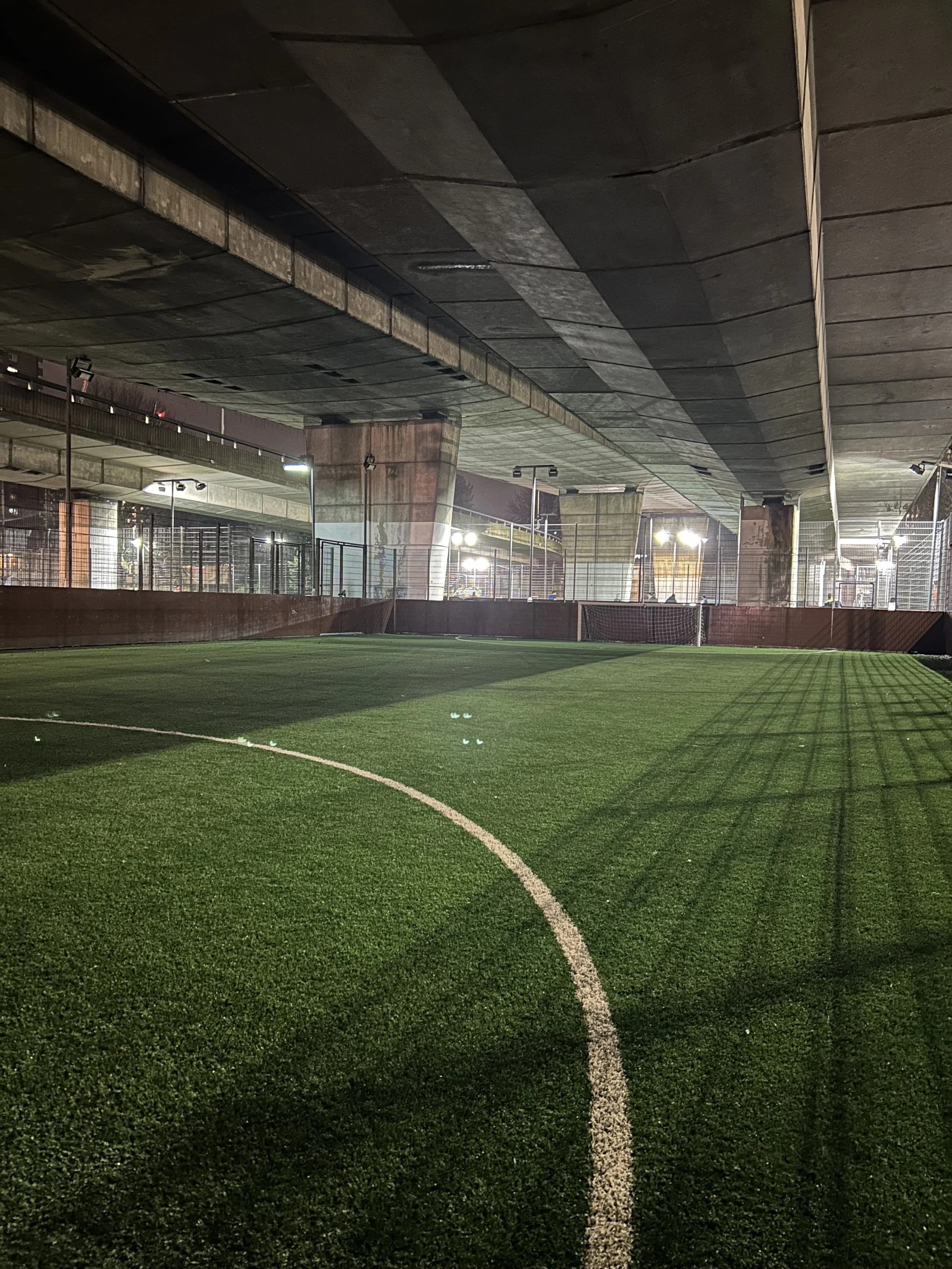 Empty artificial turf soccer field under a bridge at night, illuminated by bright lights, with a small goal at the far end and shadows from the overhead structure.