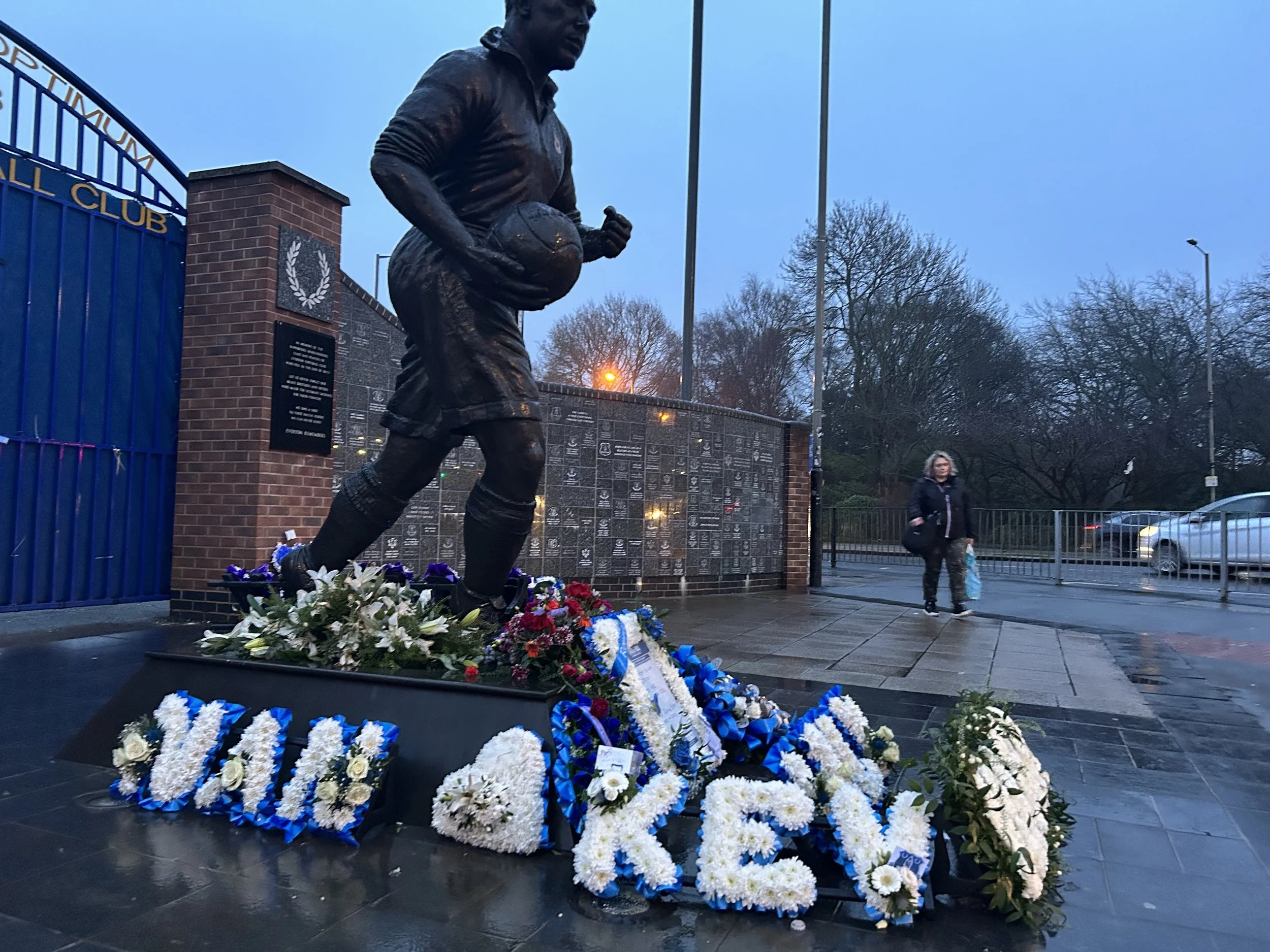 A bronze statue of a football player holding a football, surrounded by floral wreaths and arrangements, situated outside a memorial wall at a stadium entrance on a rainy evening.