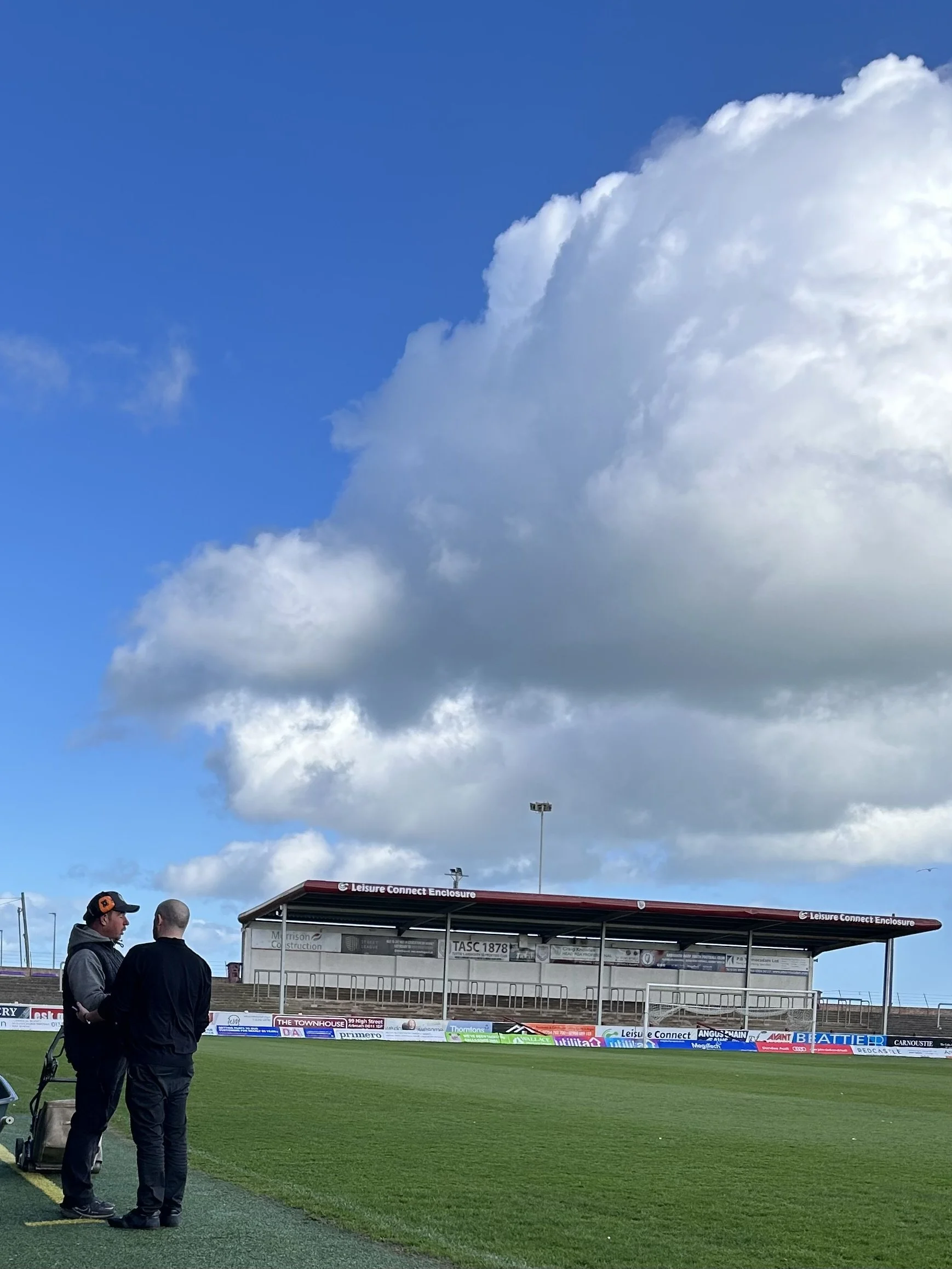 Two men standing and talking on a sports field with a seating area and large cloud in the sky.