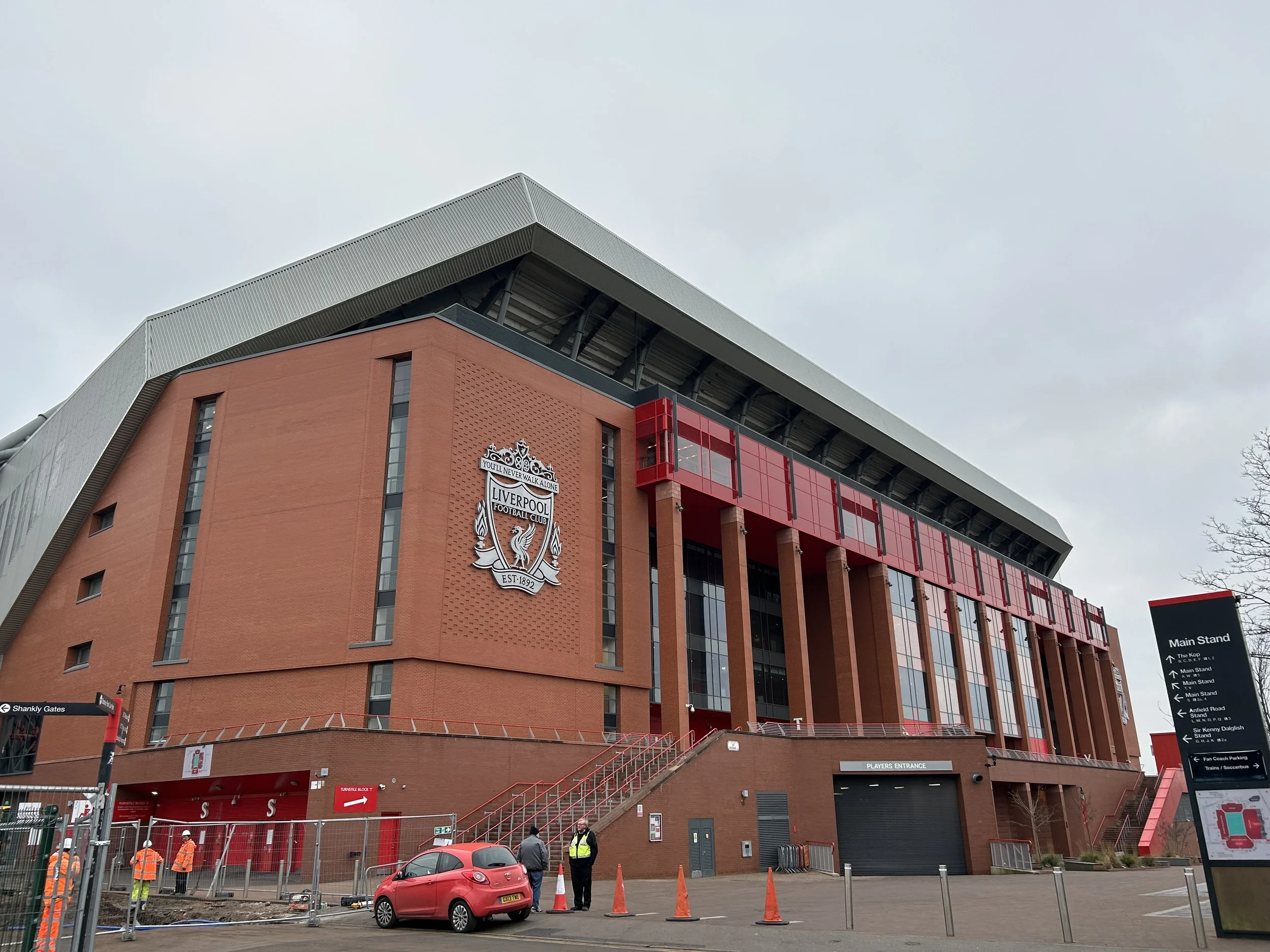 Exterior of Liverpool Football Club stadium with the club's crest, red and brick facade, and a sign pointing to the main stand and other stadium areas.