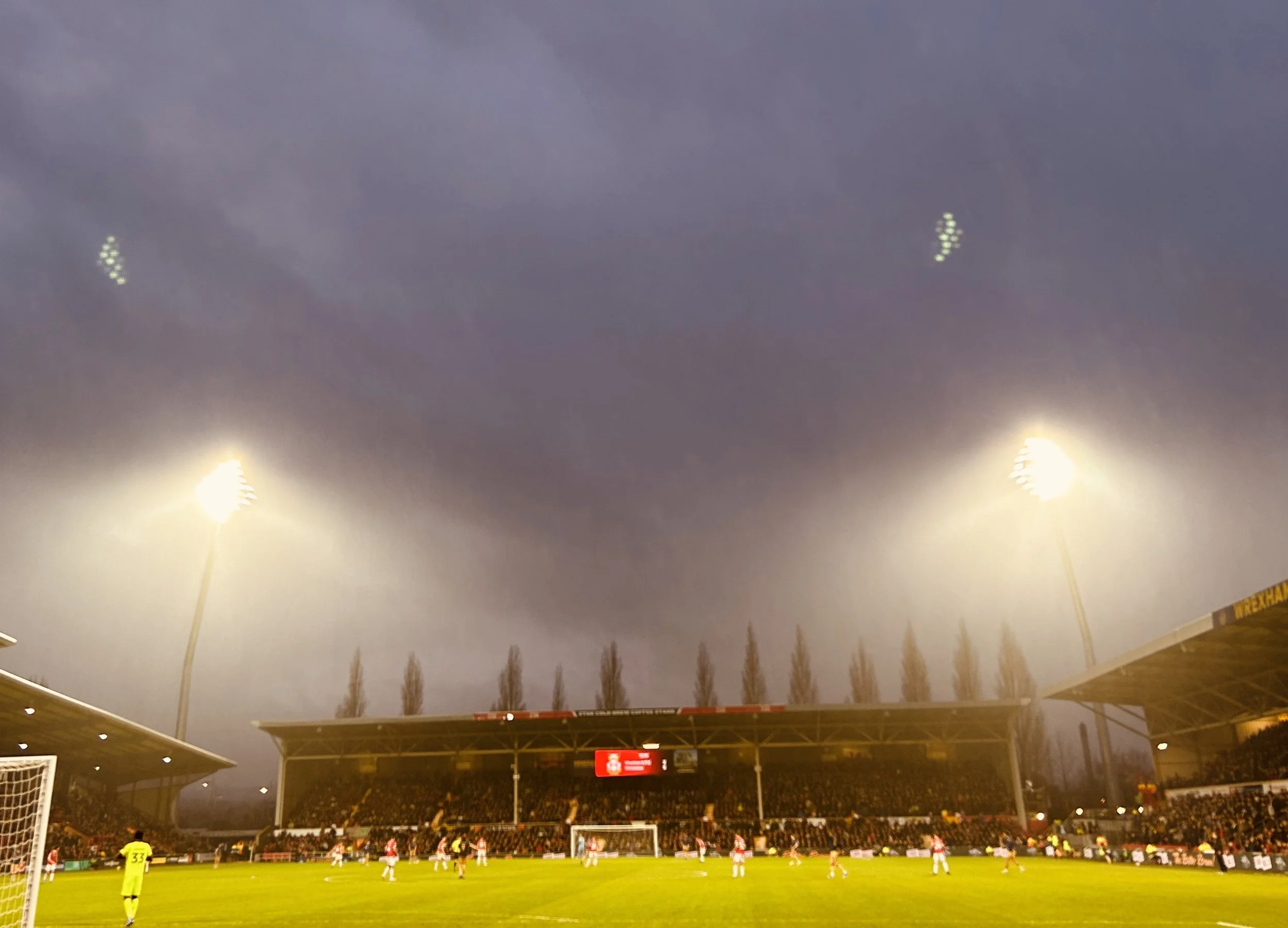 A nighttime soccer match at a stadium with players on the field and bright floodlights illuminating the scene.