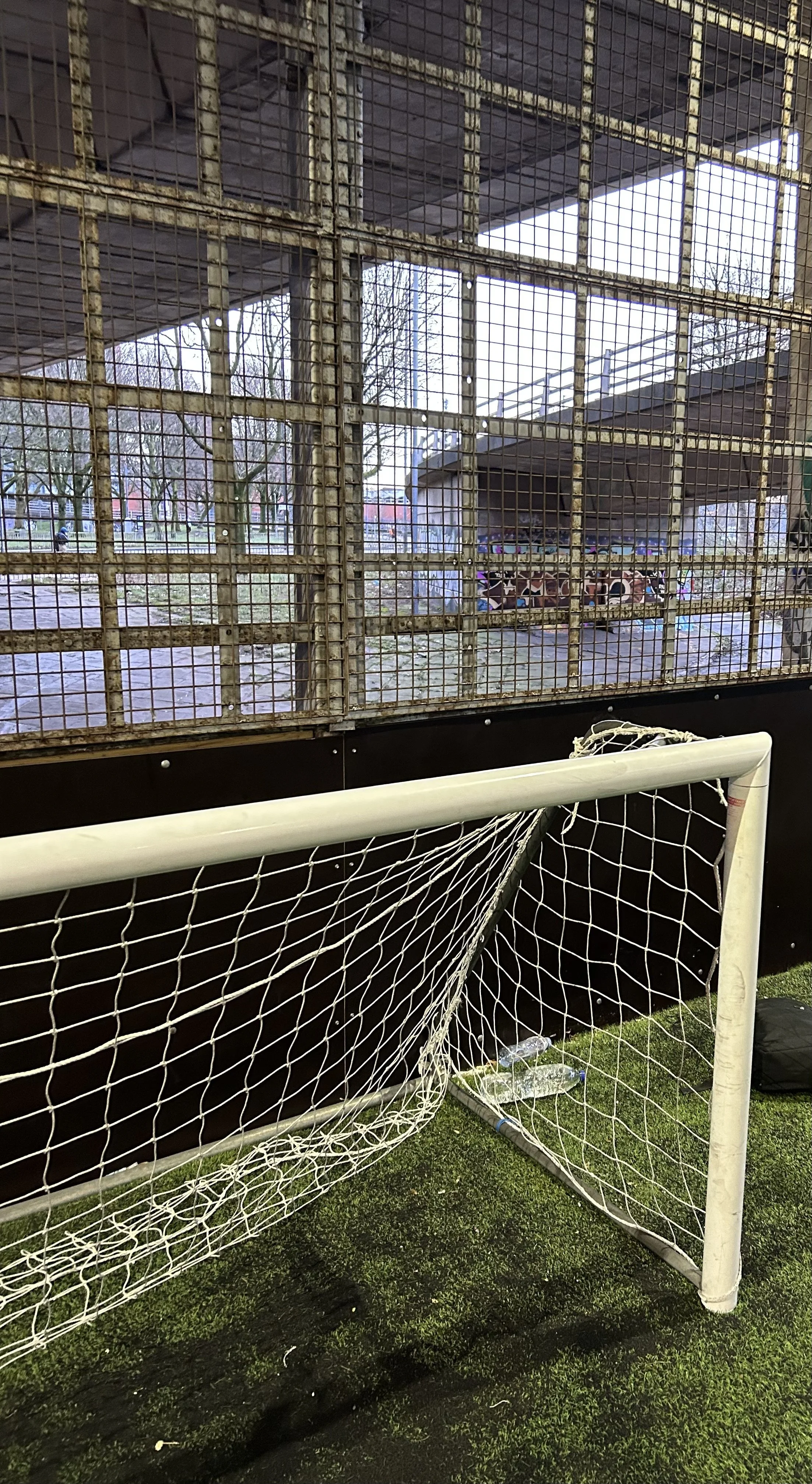 A small soccer goal on an indoor turf field with a weathered metal fence and an outdoor scene visible through it.
