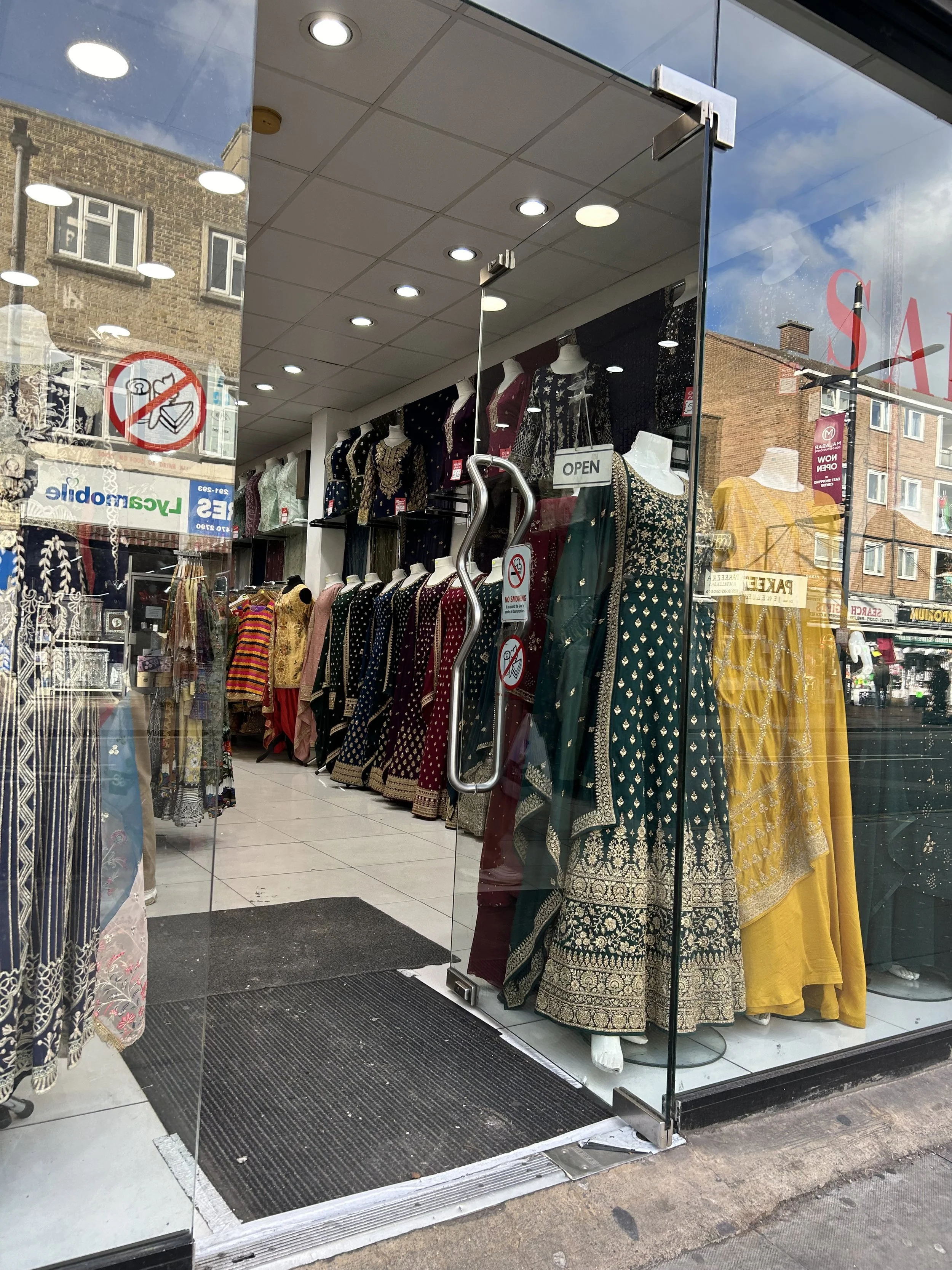 Store window displaying traditional Indian dresses in various colors and patterns, mannequins showcasing yellow, green, and other dresses, with reflections of buildings and street outside.