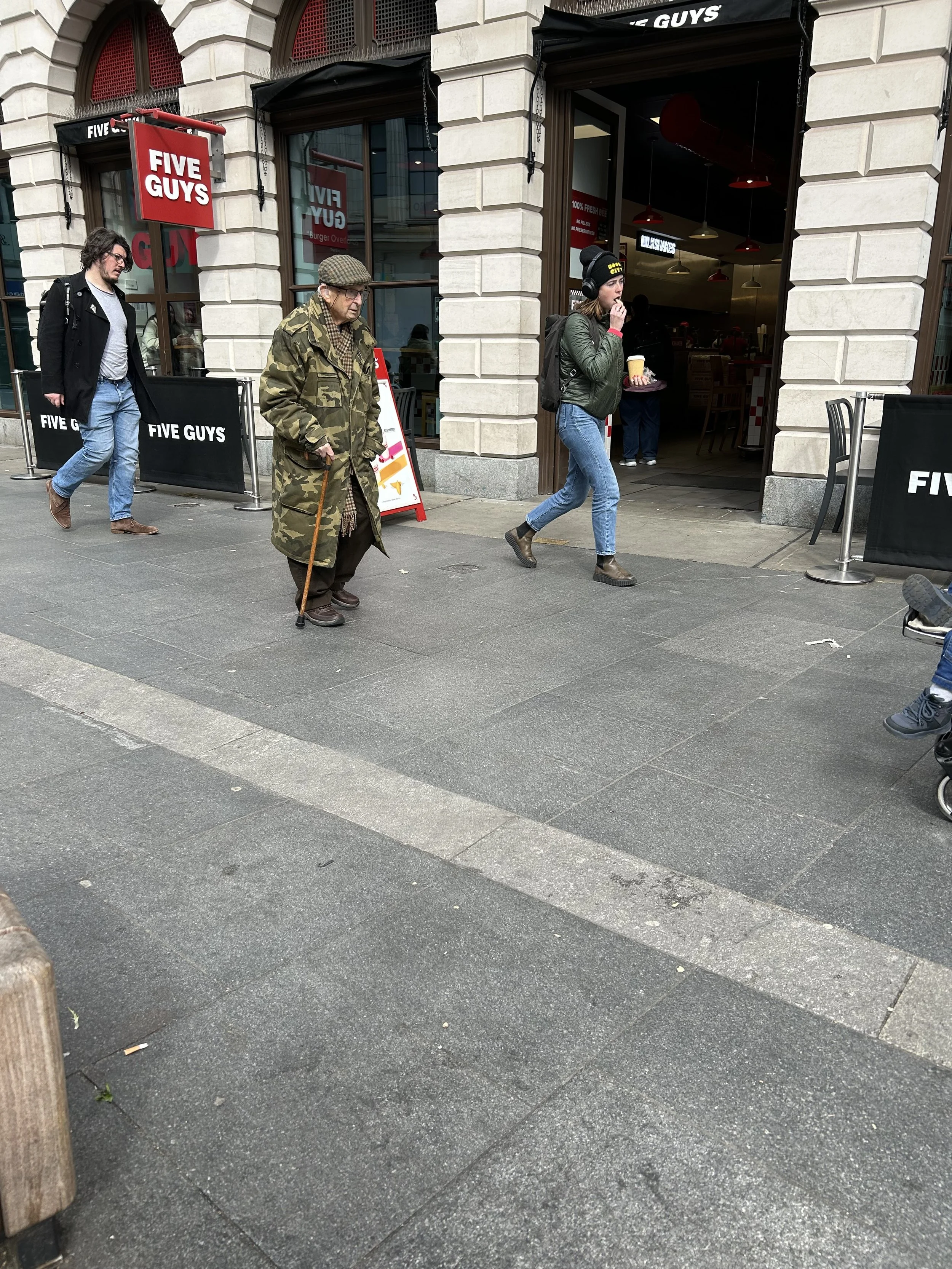 People walking outside a Five Guys restaurant, with a woman eating and drinking coffee, an elderly man with a cane wearing a camouflage coat, and another man carrying a backpack.