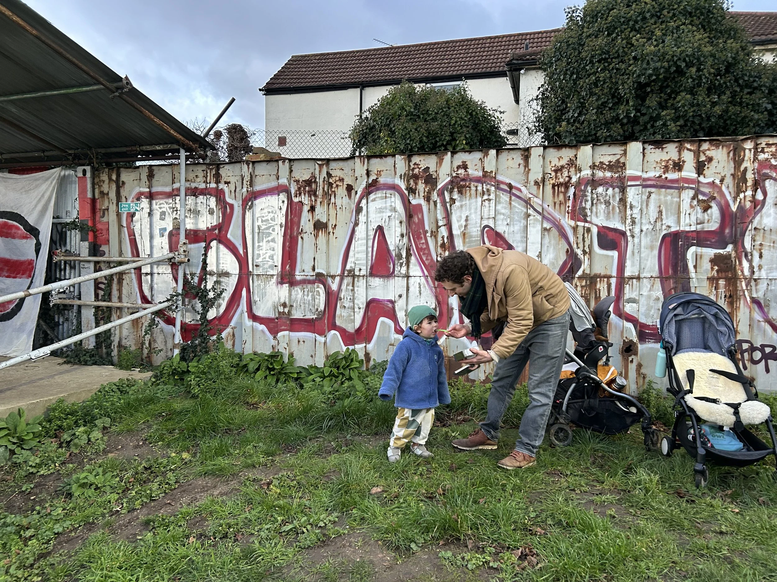 A man and a young boy in front of a rusty metal fence with graffiti. The man is feeding or talking to the boy, who wears a green hat and colorful pants. There are two strollers next to them on the grass, and the scene is outdoors with buildings in th