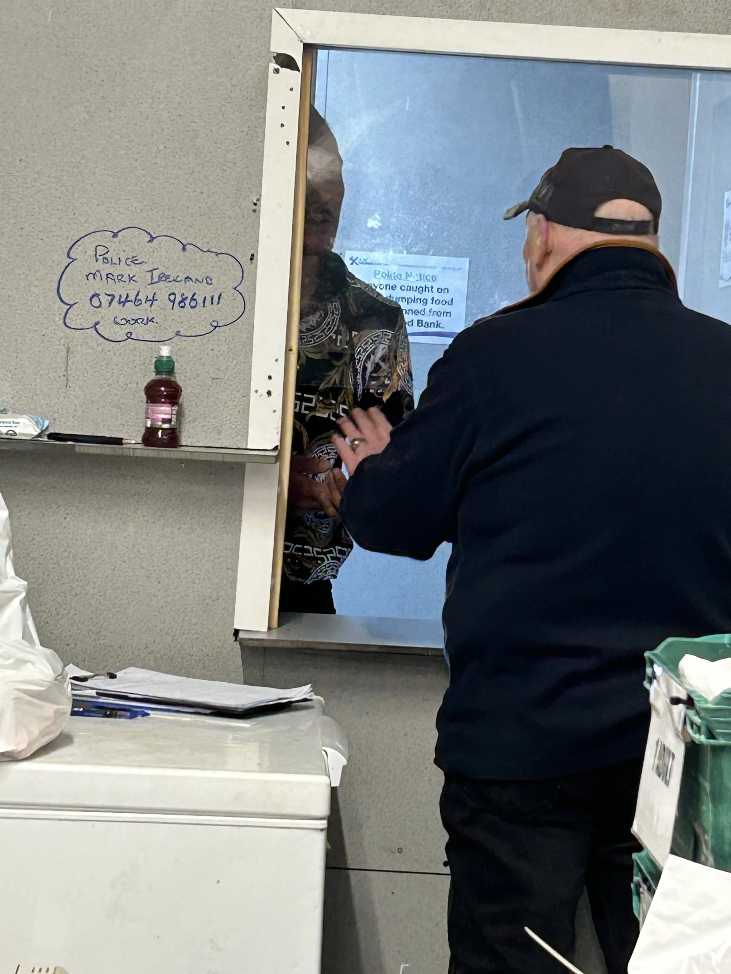 A man in a black cap and jacket is talking to a woman behind a service window, with a whiteboard and papers in the foreground.