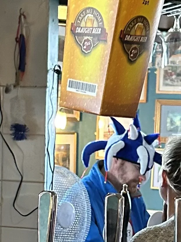 A man wearing a Sonic the Hedgehog hat at a bar or restaurant, with a yellow box labeled "Take Me To Ddaughter Jea" hanging above him in a decorated setting.