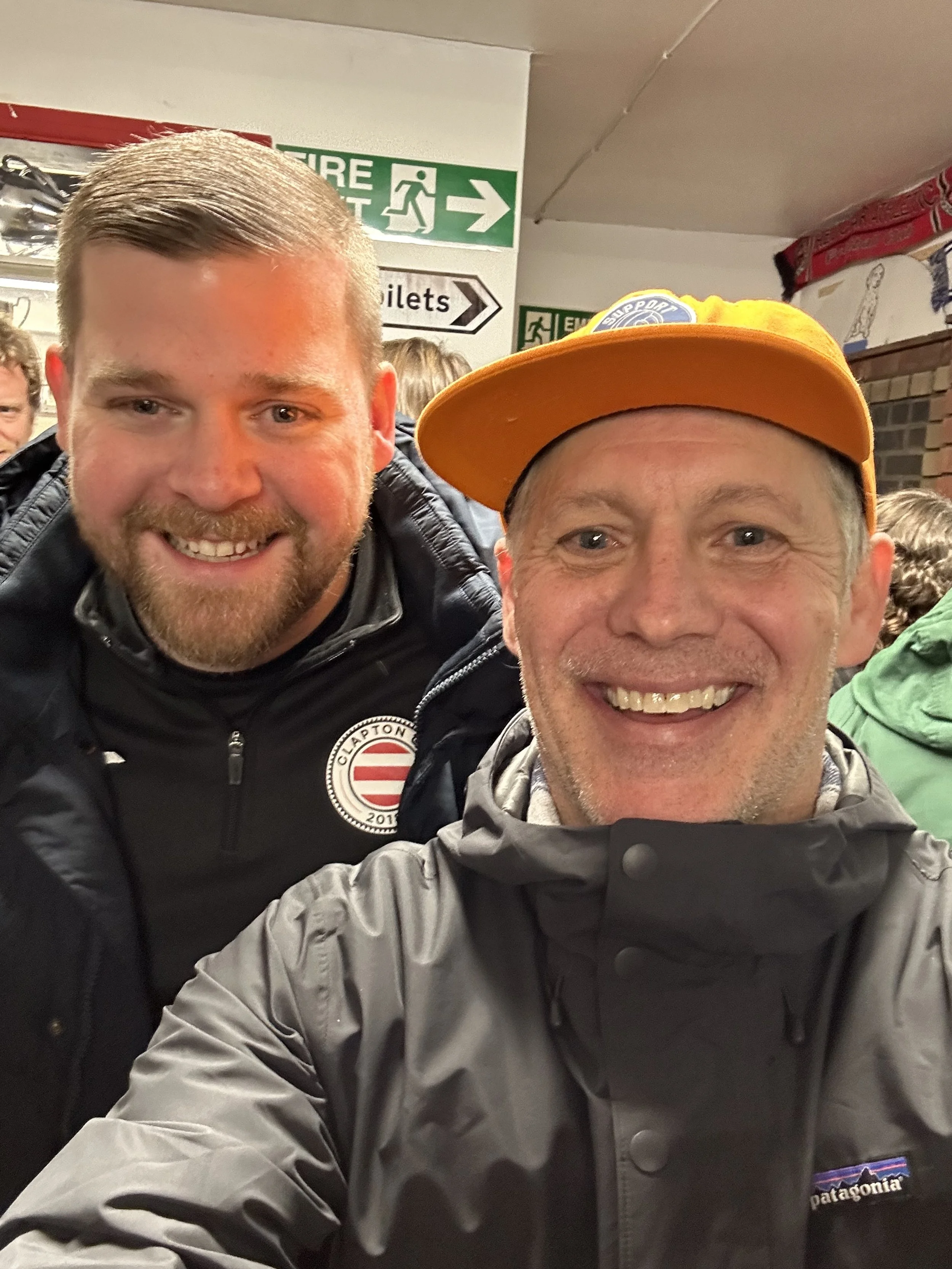 Two smiling men taking a selfie indoors, with signs for fire exit and toilets visible in the background.