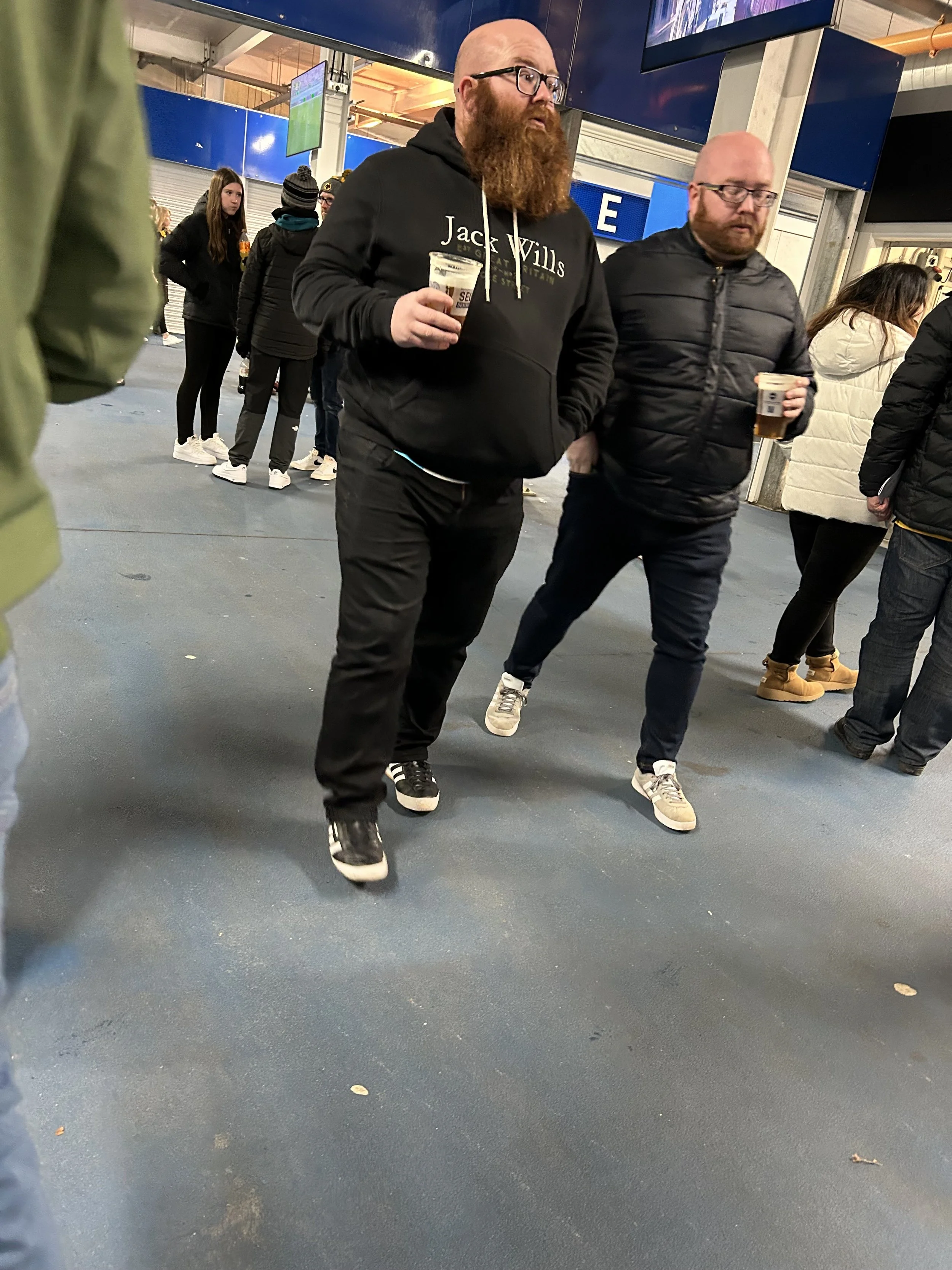 Two men walking in a crowded indoor space, each holding a plastic cup with dark beverage. The man on the left has a long red beard, glasses, black hoodie with 'Jack Wills' printed on it, and black pants. The man on the right is bald, has glasses, and