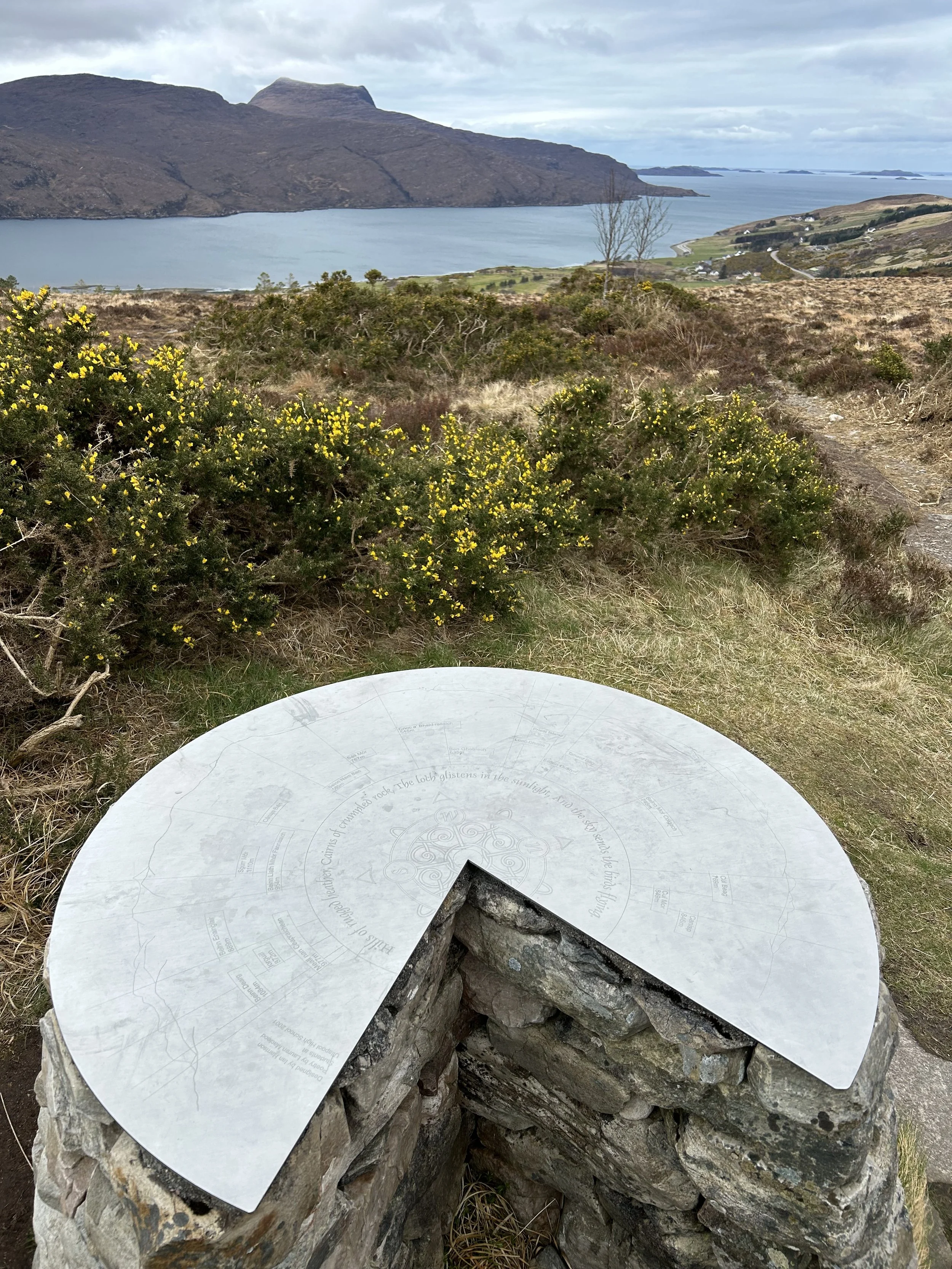 A circular informational sign mounted on a stone pedestal with a scenic landscape of mountains, water, and yellow flowering bushes in the background.