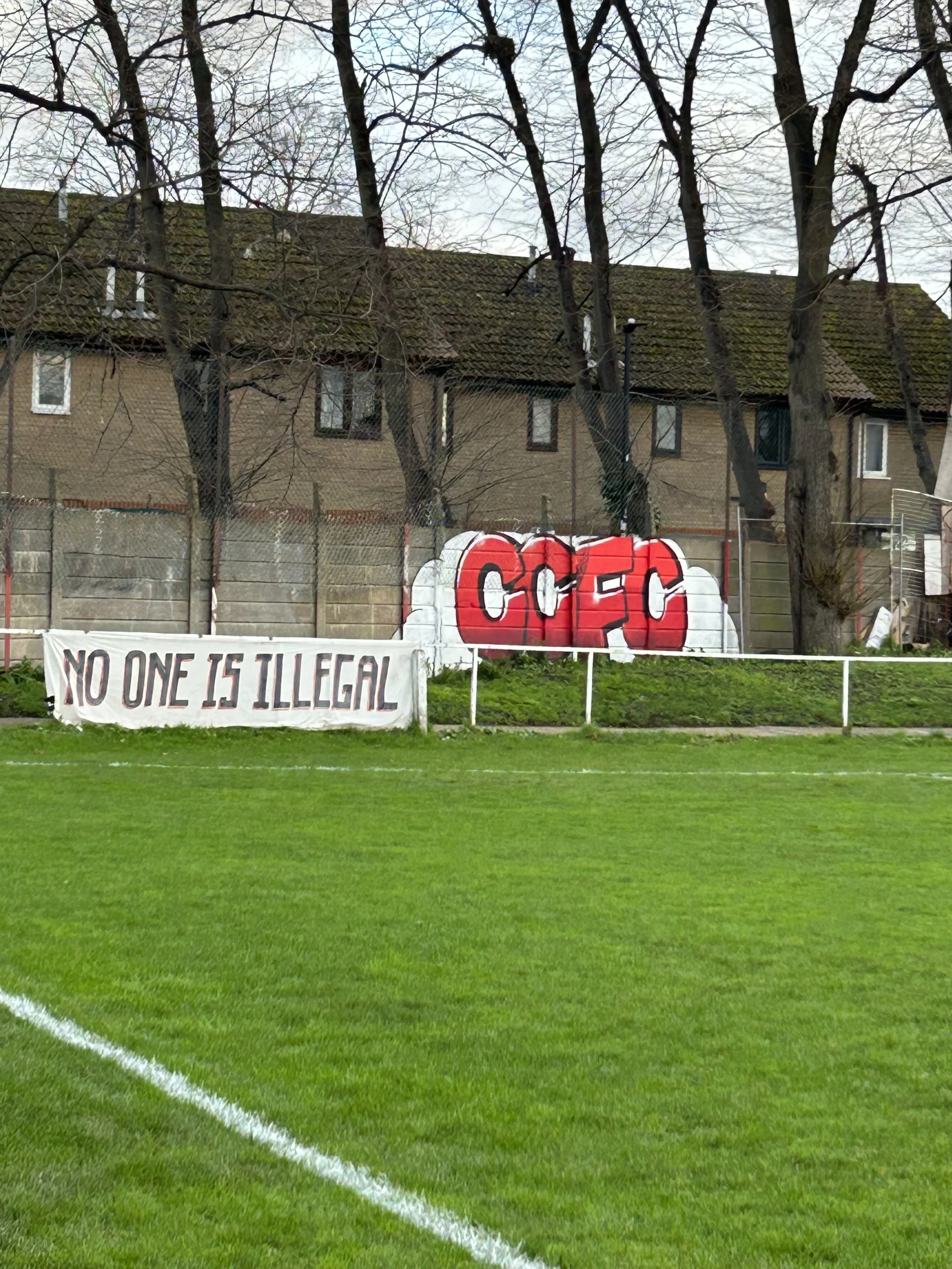 A graffiti artwork spelling 'CFC' in red and white spray paint on a wall behind a fence, with a banner reading 'No one is illegal' in front. The setting is a grassy field with white boundary lines, and there are leafless trees and residential buildin