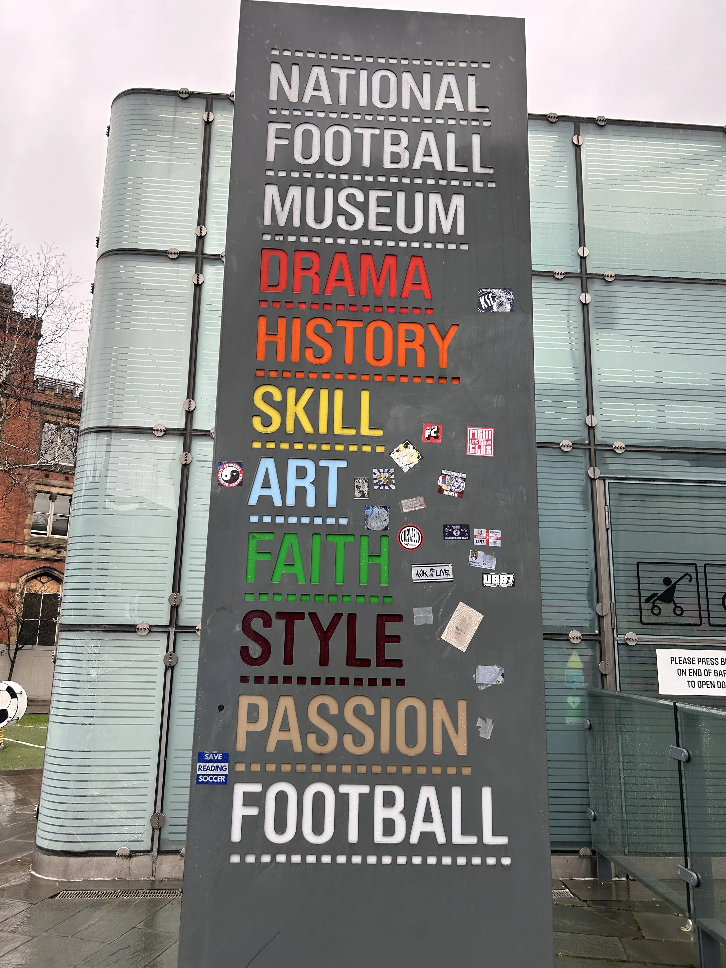 A large, vertical sign outside the National Football Museum displays various words in different colors, including 'DramA,' 'History,' 'Skill,' 'Art,' 'Faith,' 'Style,' 'Passion,' and 'Football.' The background shows modern glass building architecture
