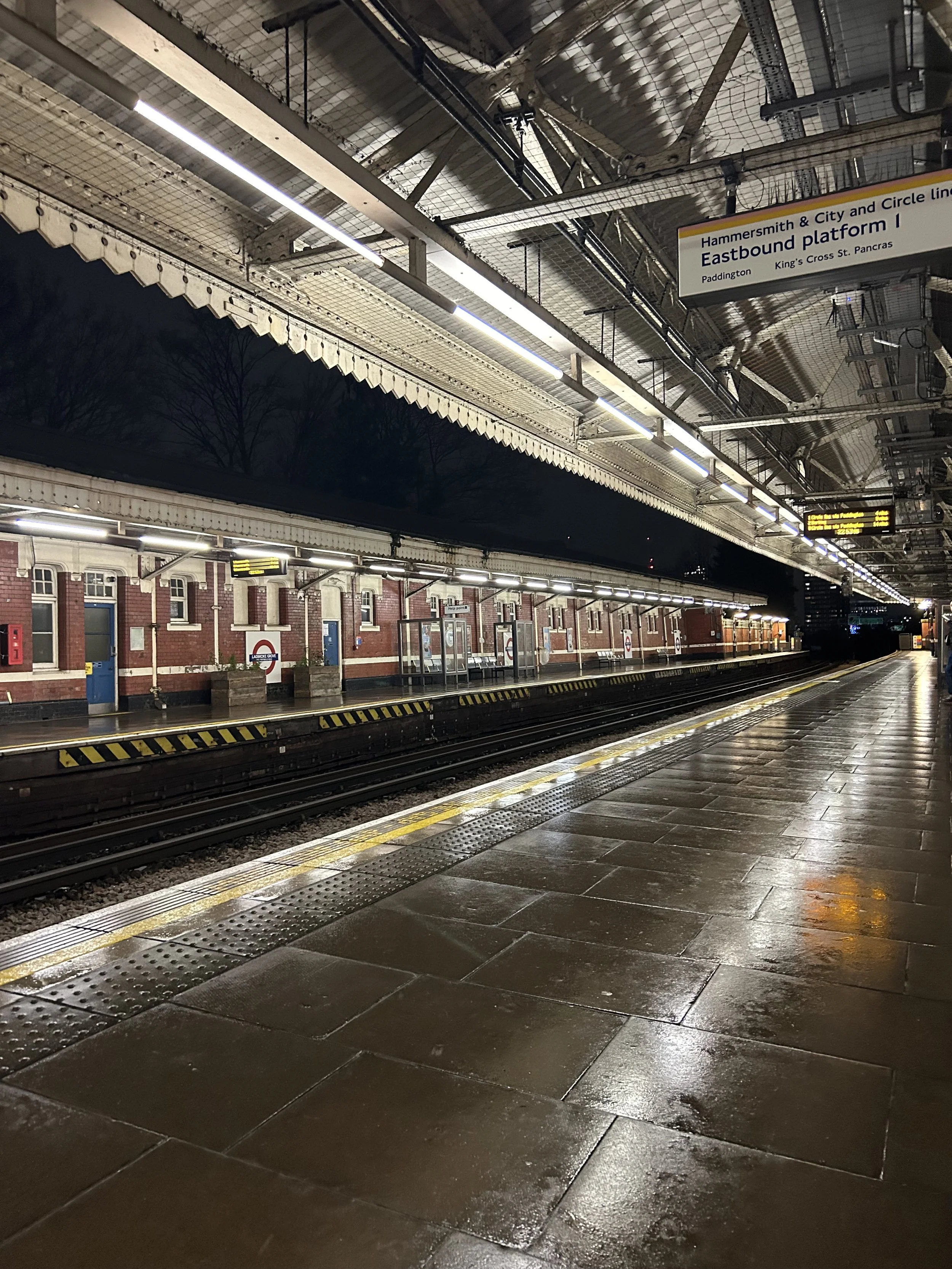 Empty London underground train station platform at night, wet floor, red brick buildings across the tracks, overhead signs for Eastbound platform, and dark sky with trees in the background.