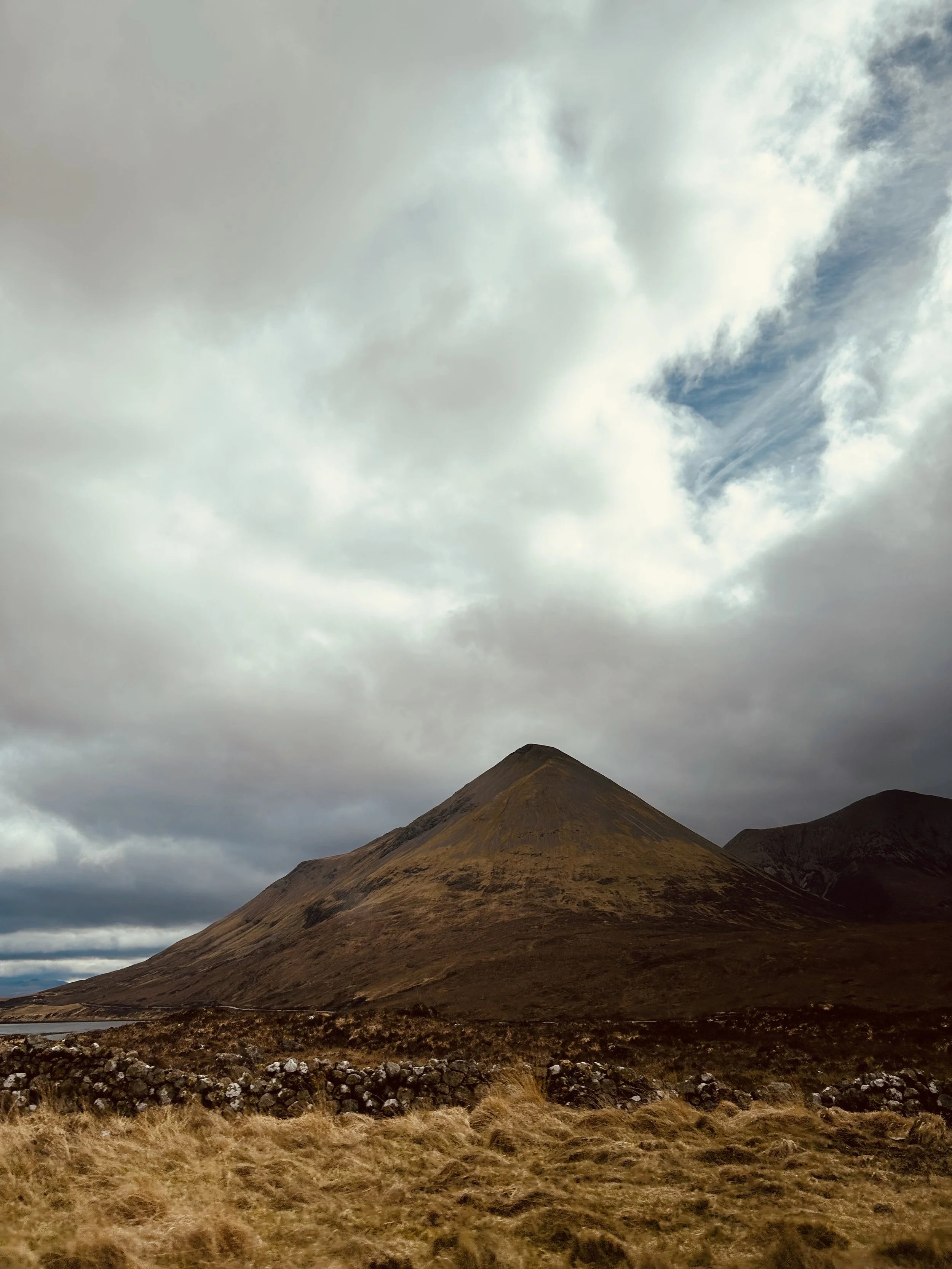 A mountain with a rounded peak surrounded by grassy and rocky terrain under a cloudy sky with patches of blue.