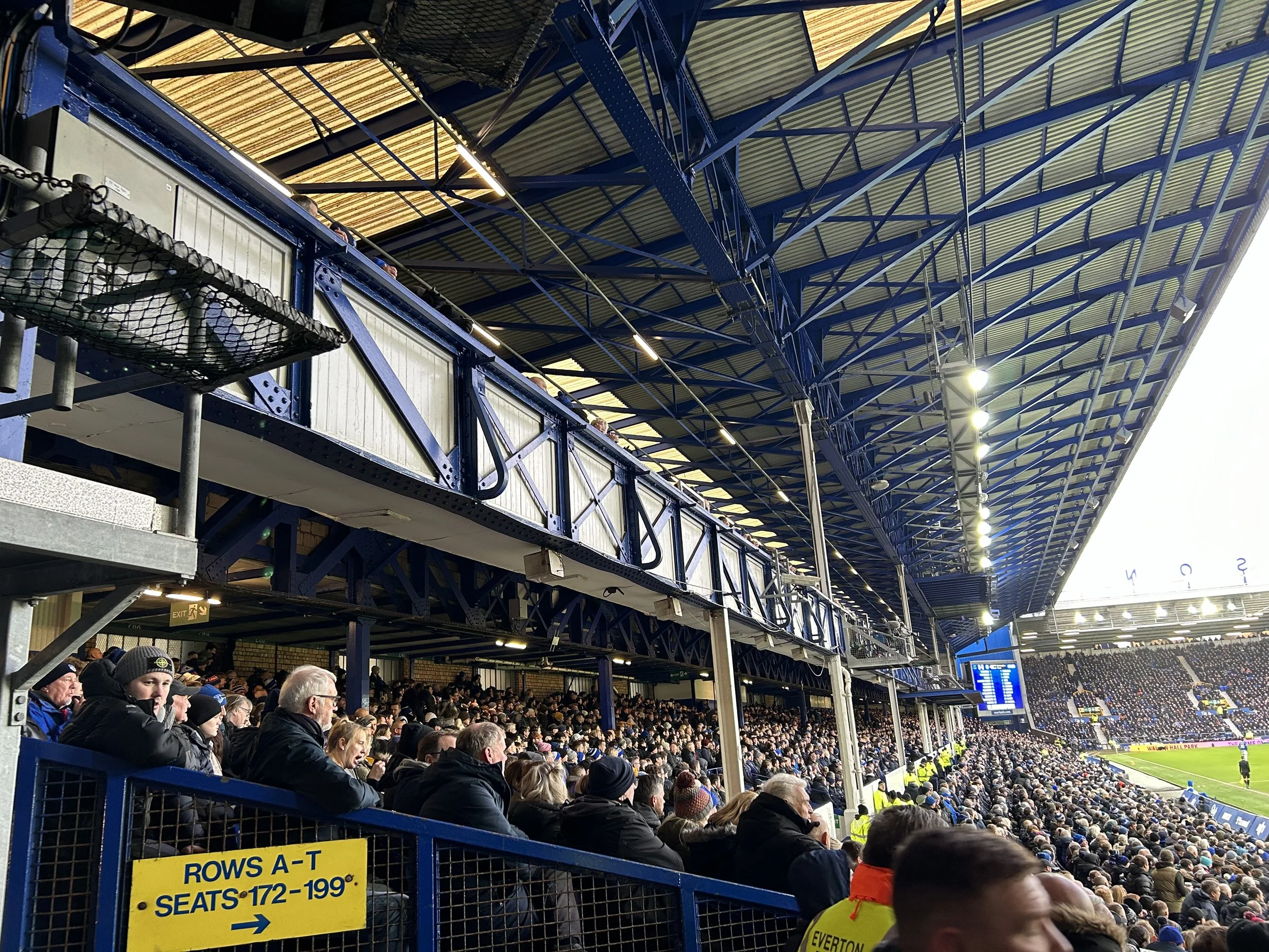 Crowd of people seated in a stadium watching a sports game, with a sign indicating rows A-T and seats 172-199.