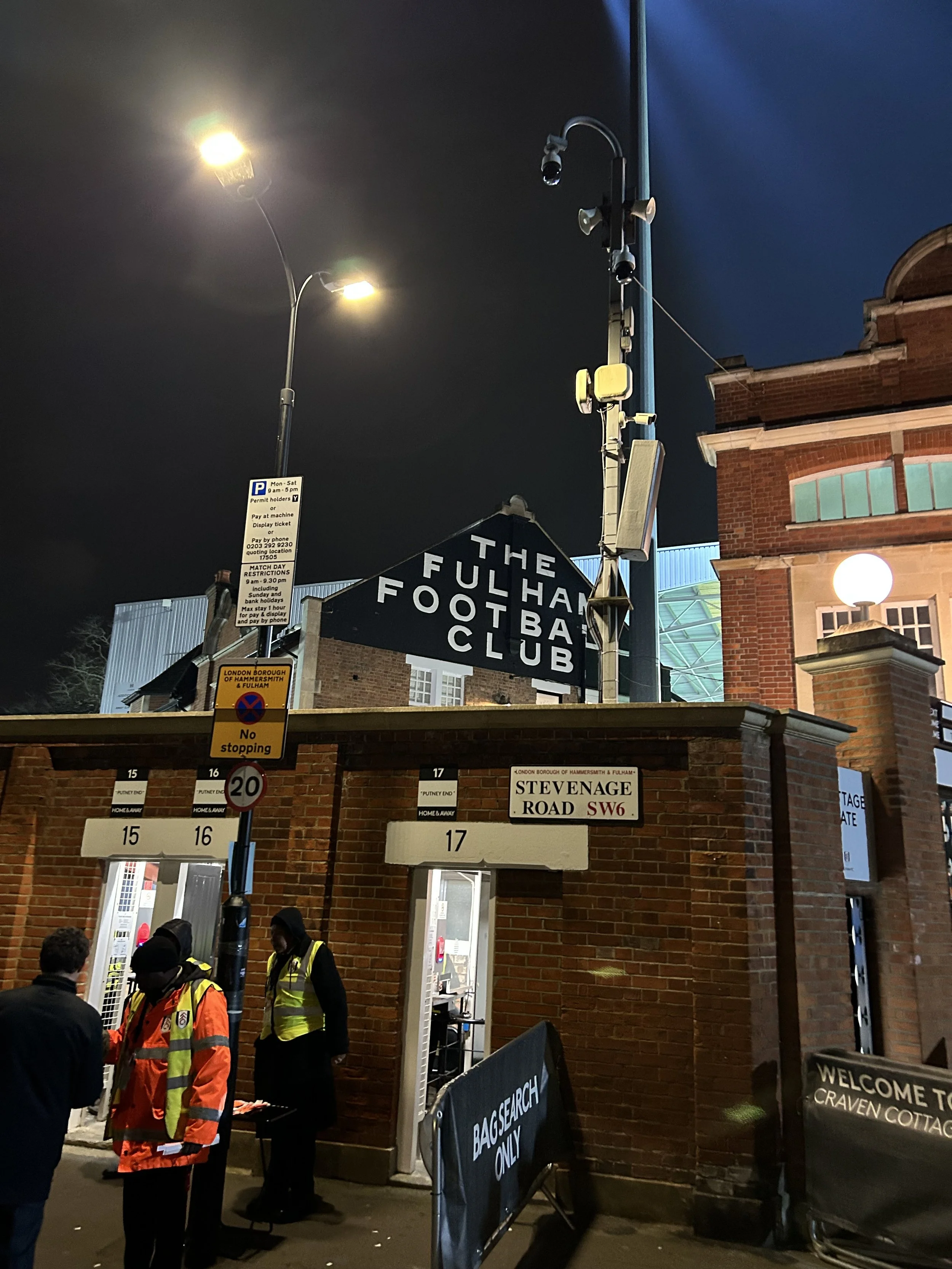 Nighttime scene outside The Fulham Football Club entrance with staff and security personnel, brick building, signage including 'Stevenage Road SW6,' and streetlights.