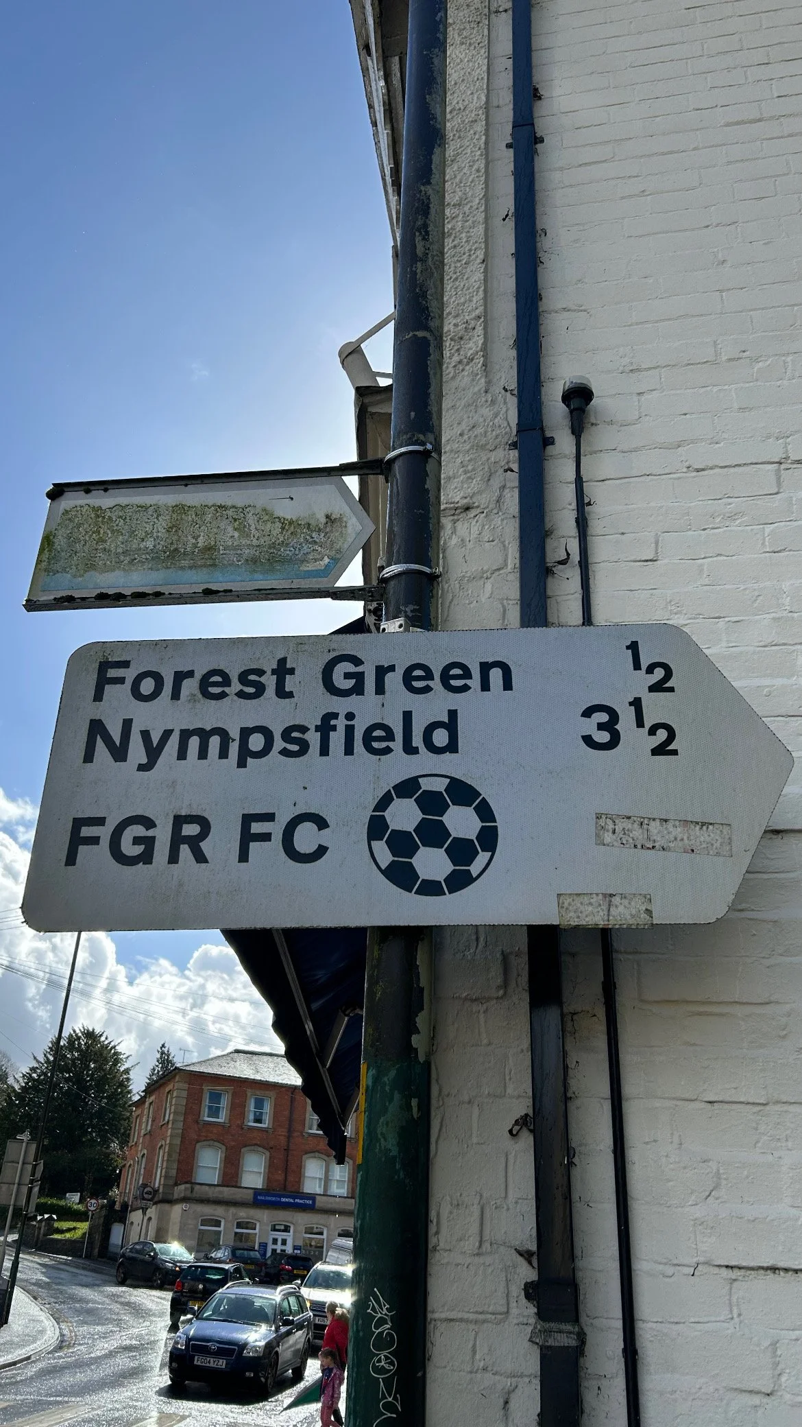 Street sign showing directions to Forest Green, Nympsfield, and FGR FC with distances in miles, mounted on a pillar next to a white brick building, with vehicles and buildings visible in the background.