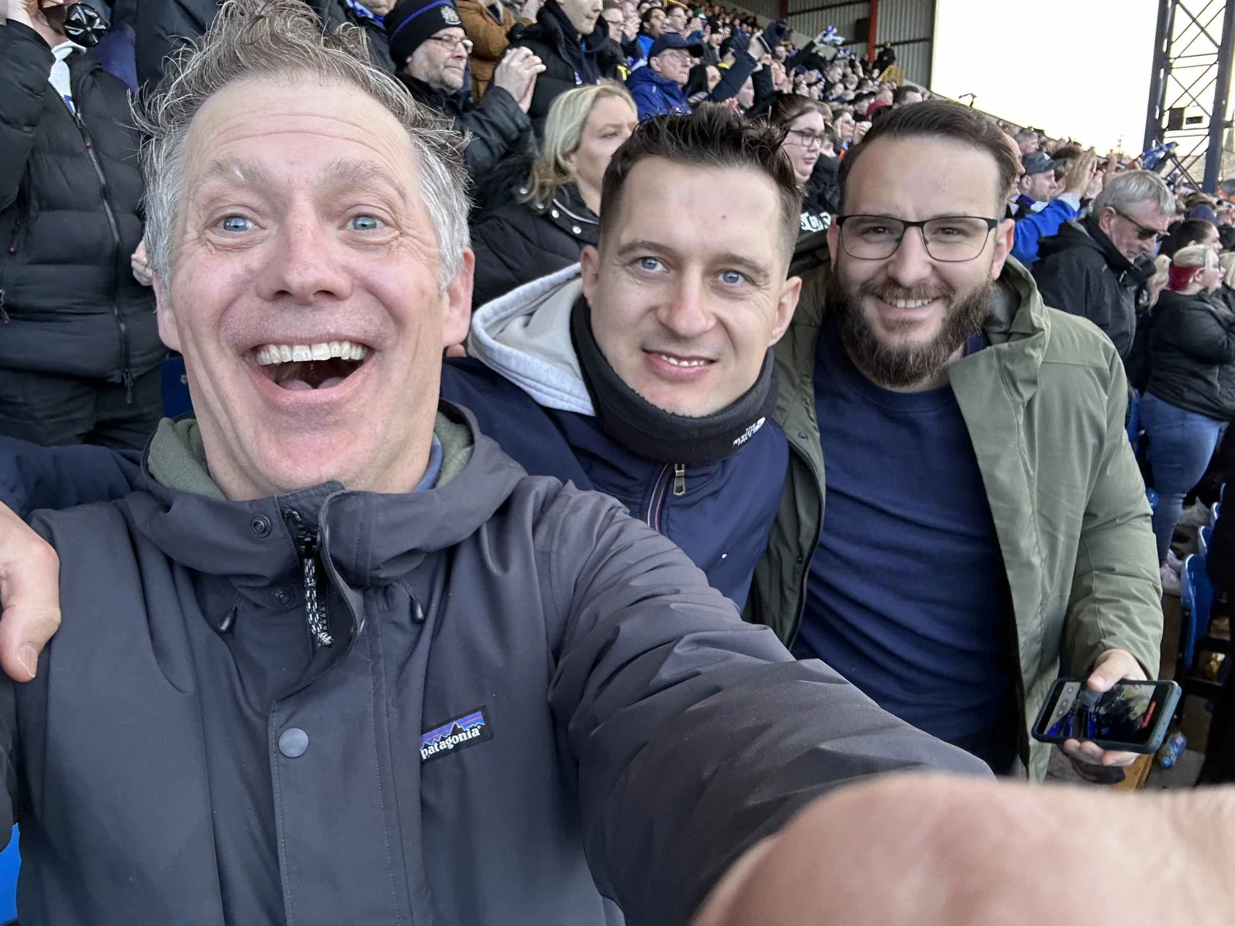 Three men taking a selfie at a crowded outdoor event with a large audience in the background.