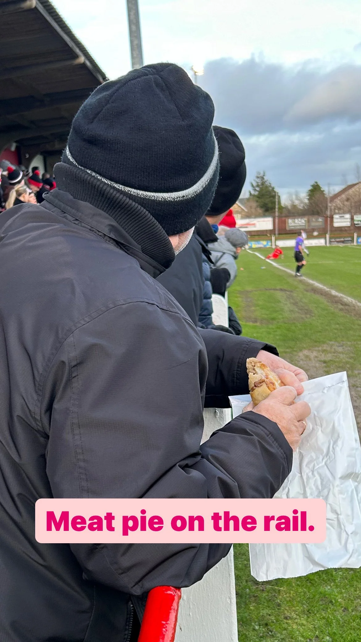 A person wearing a black beanie and black jacket holding a meat pie in a white paper bag, standing at a sports field with others in the background, some watching a game.