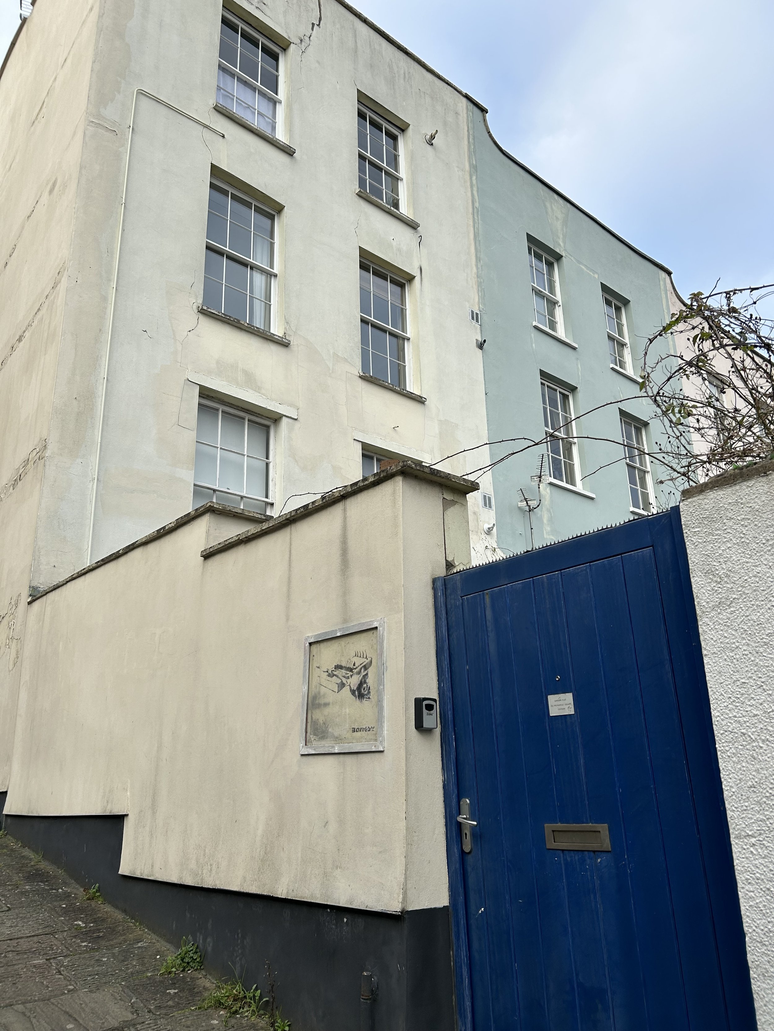 A multi-story old building with peeling paint, multiple windows, and a blue gate in front. There is a poster on the wall and some wires, with a cloudy sky above.