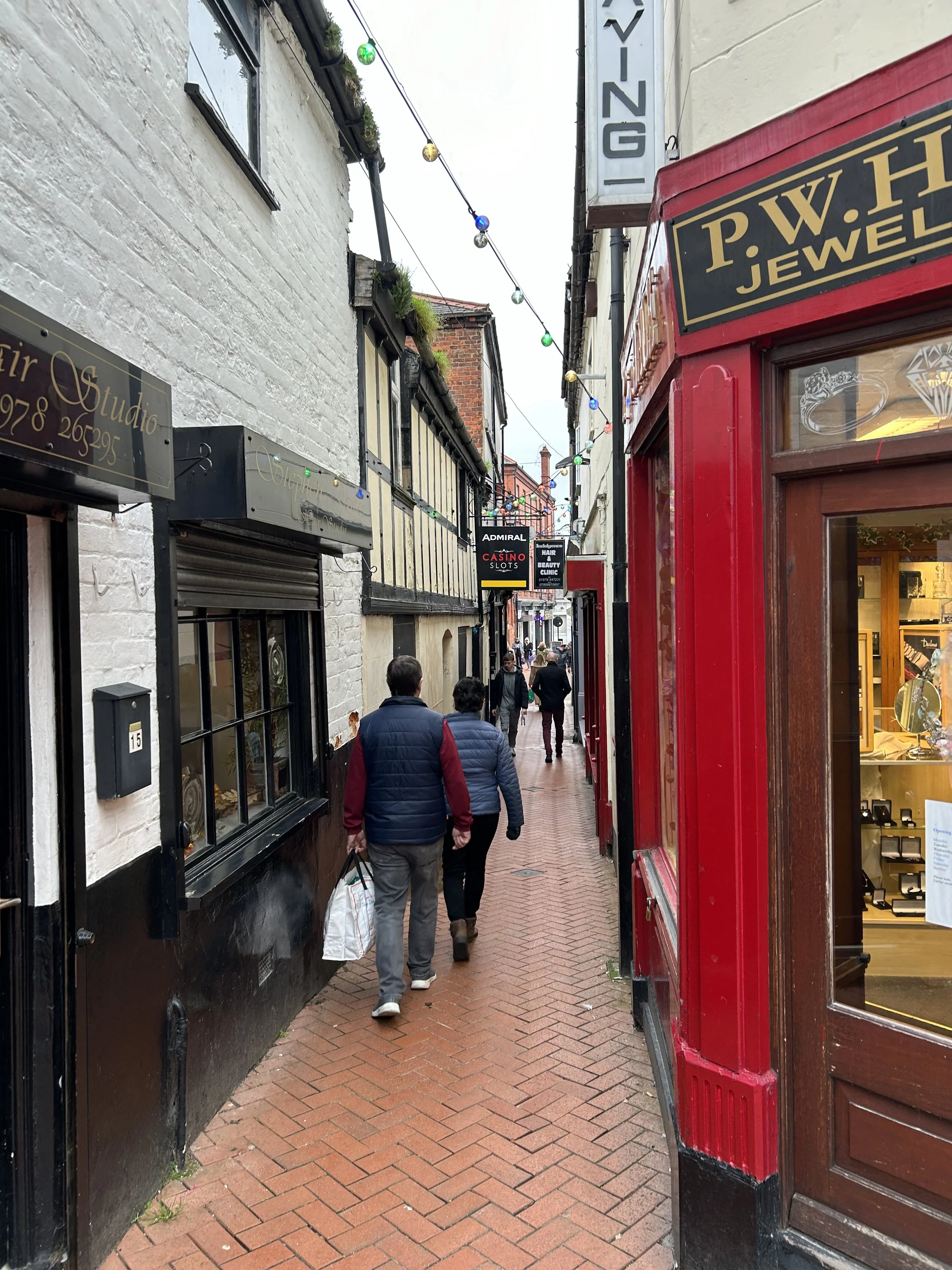People walking on a narrow brick sidewalk between storefronts under string lights in an urban area.