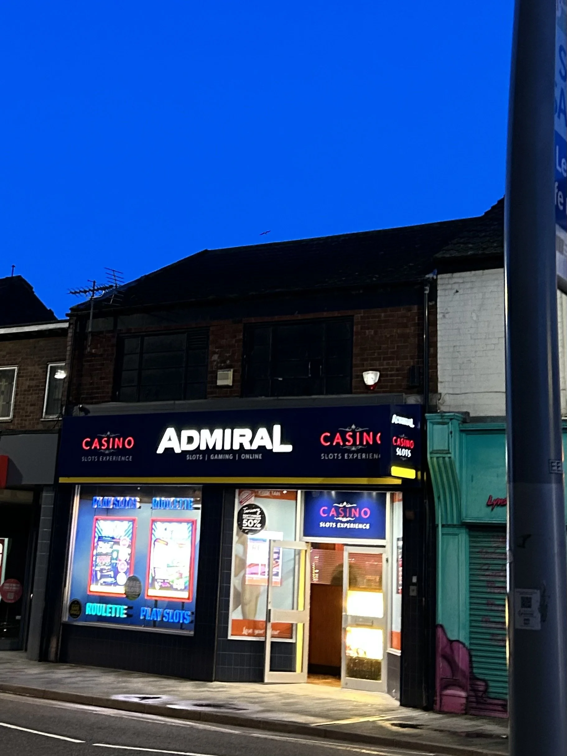 Night view of a betting shop called Admiral with bright neon signs advertising casino games, slots, roulette, and online gambling. The store has a black and blue exterior with signs in red, white, and blue, and there is a closed storefront with a pai