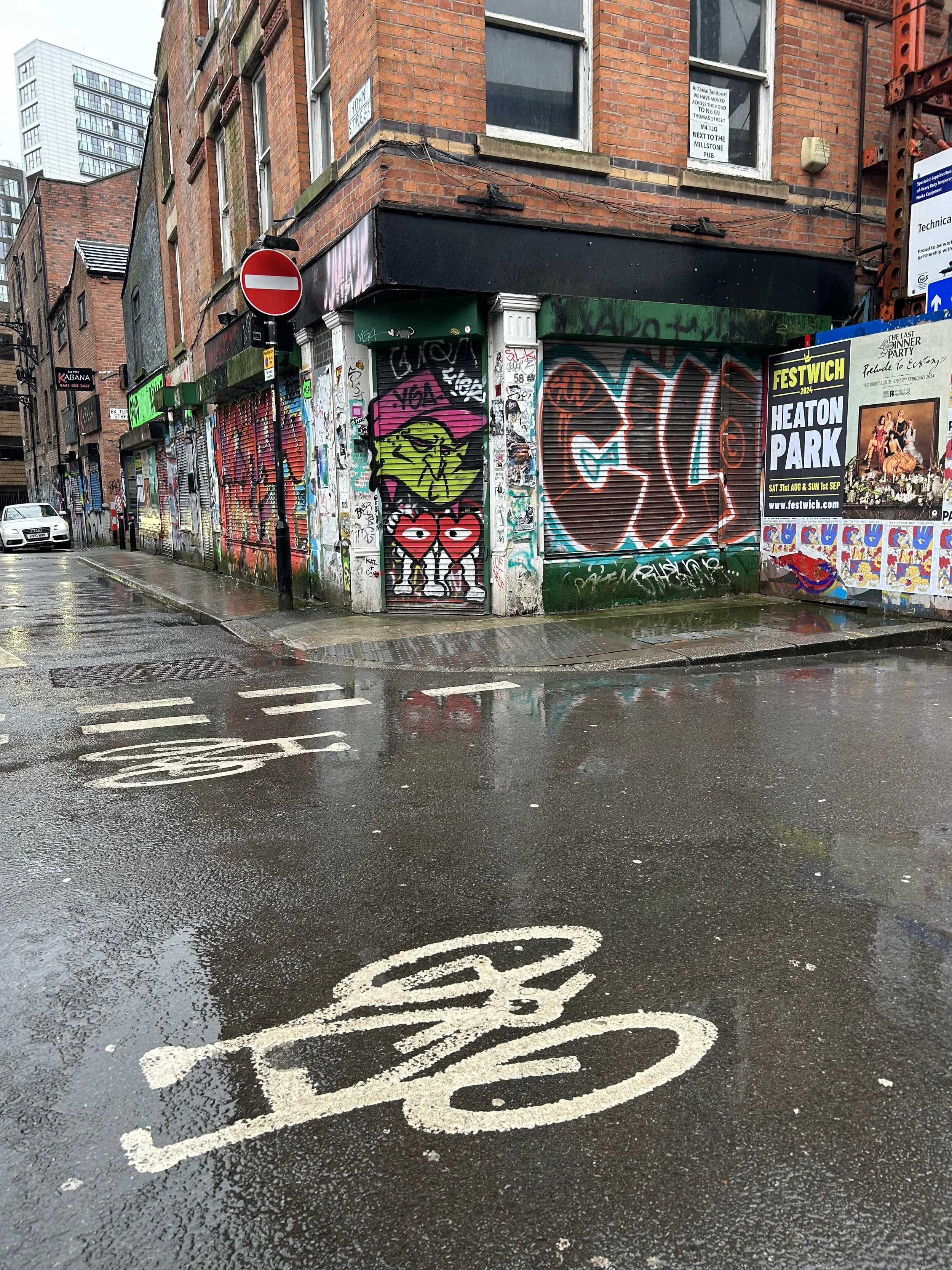 A rainy street corner with graffiti-covered storefronts, including a cartoon character with sunglasses and heart-shaped glasses on a metal shutter. The street has puddles reflecting the graffiti and buildings, with a 