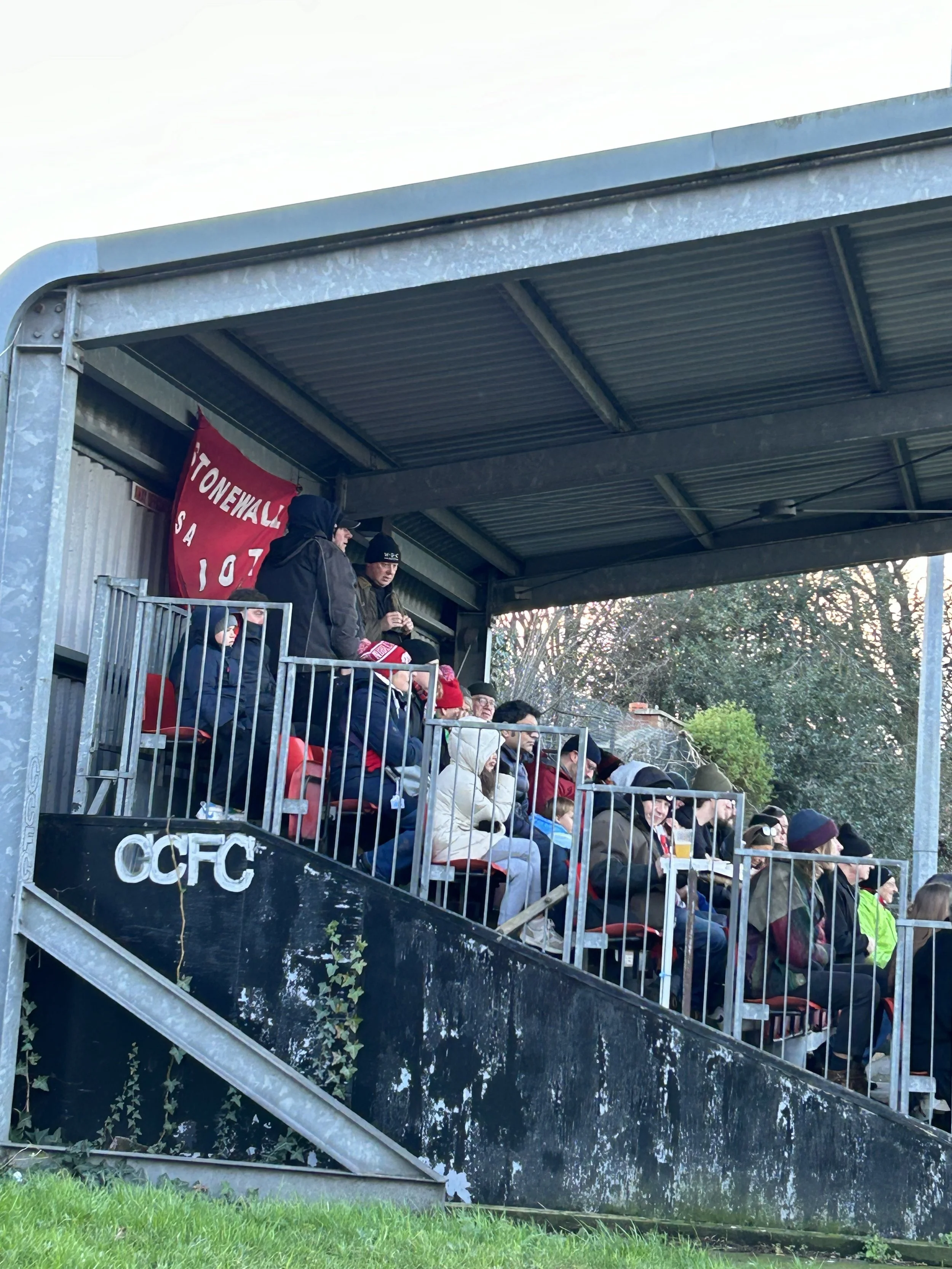 People seated under a metal covered stand, watching a sports event, with a red banner in the background that reads 'Stonewall' and '197'.