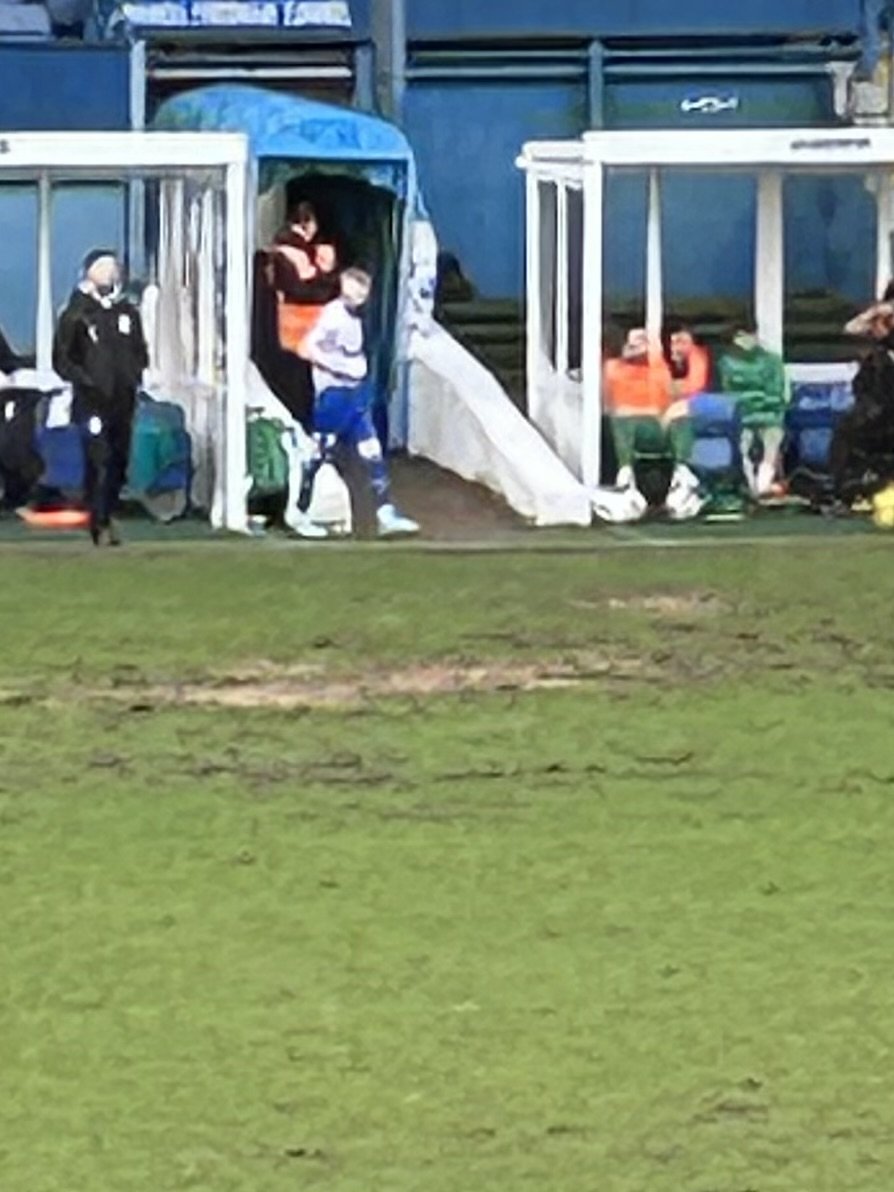 People sitting and standing near a sports field sideline, some wearing masks and sports gear, with a few children and adults, and a small structure with benches or seating areas.