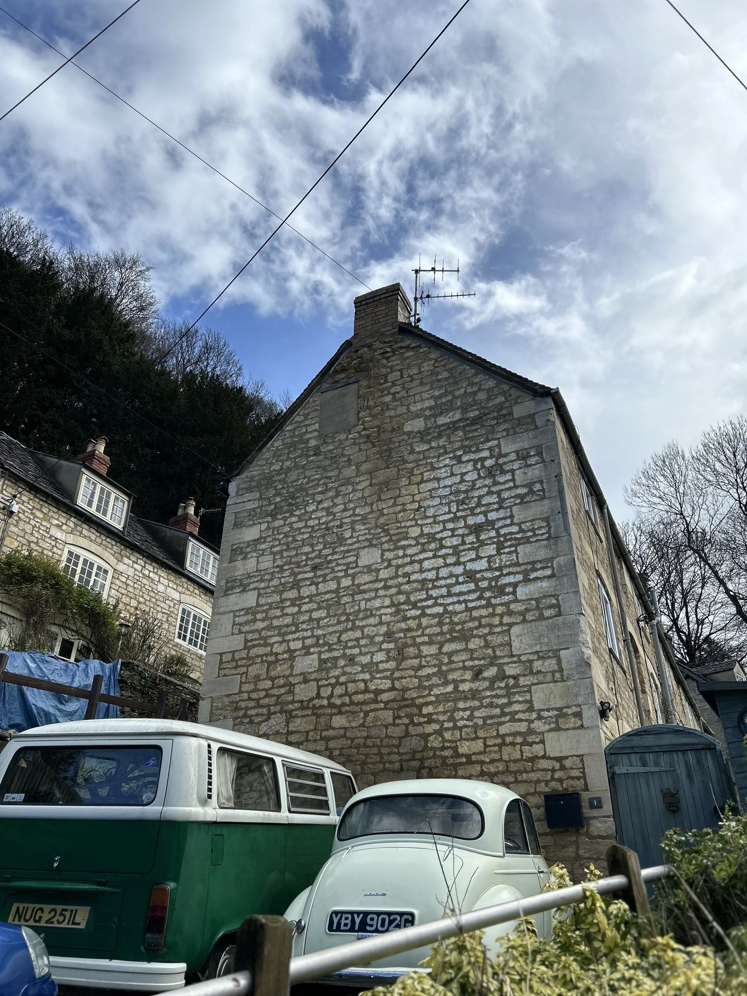 A stone building with a chimney and TV antenna, next to two vintage cars parked outside, on a cloudy day.