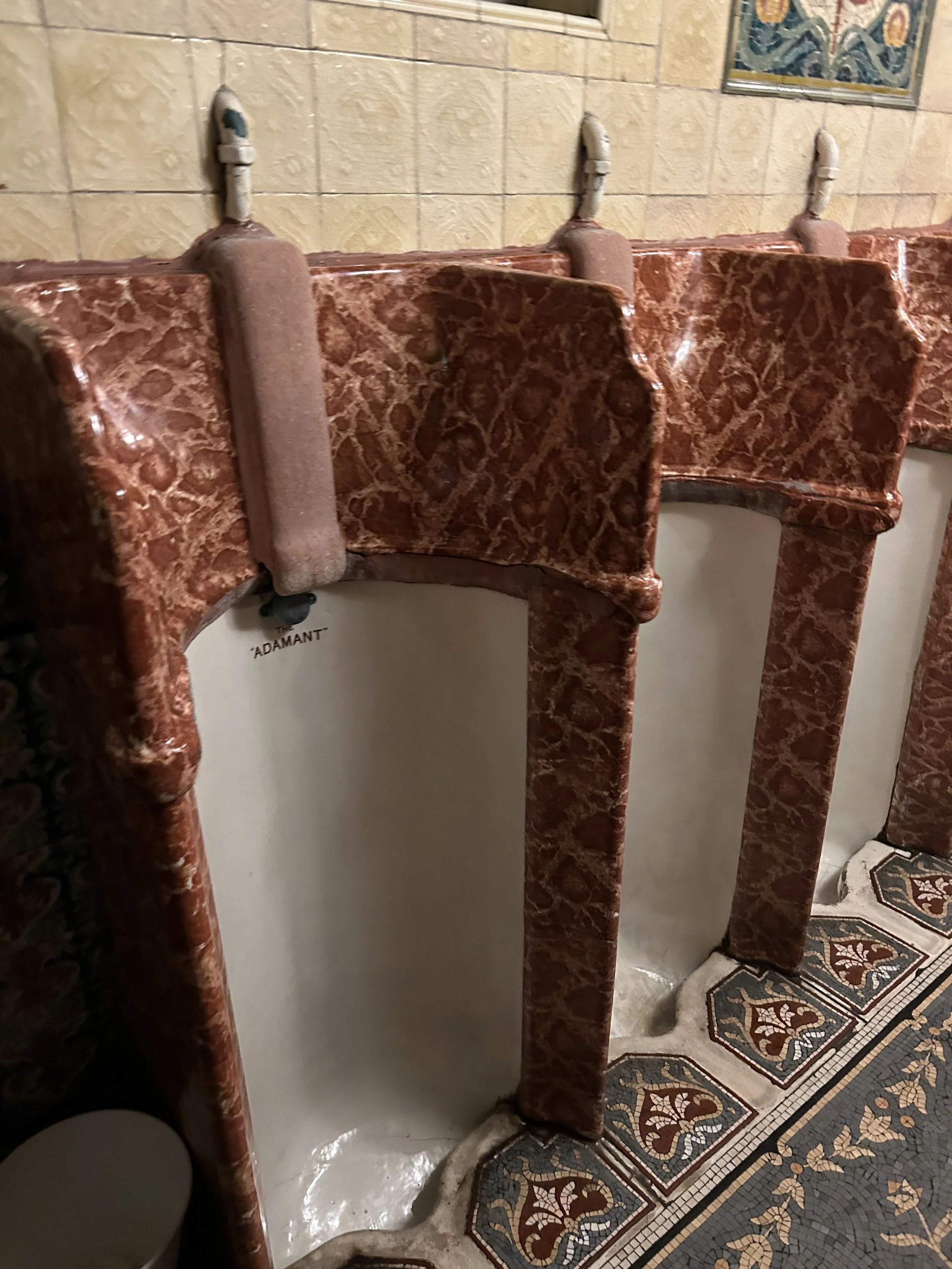 Row of vintage marble urinals with pinkish-brown marble surfaces and beige tiled wall in background, located in a public restroom.