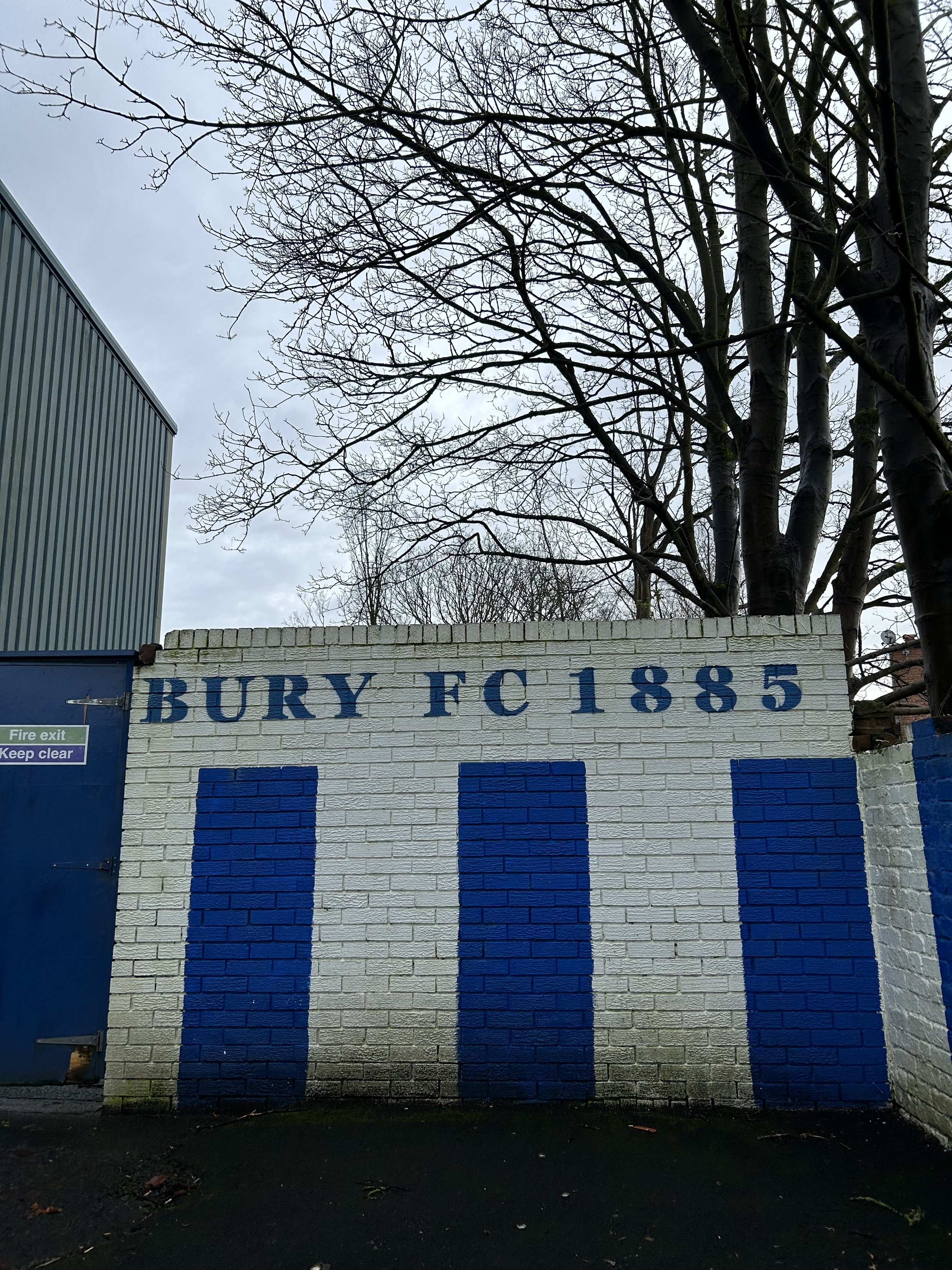 A brick wall with painted blue text reading 'BURY FC 1885' and three vertical bars below the text. There is a tree with leafless branches above the wall and a blue door on the left side with a 'Fire exit Keep clear' sign.