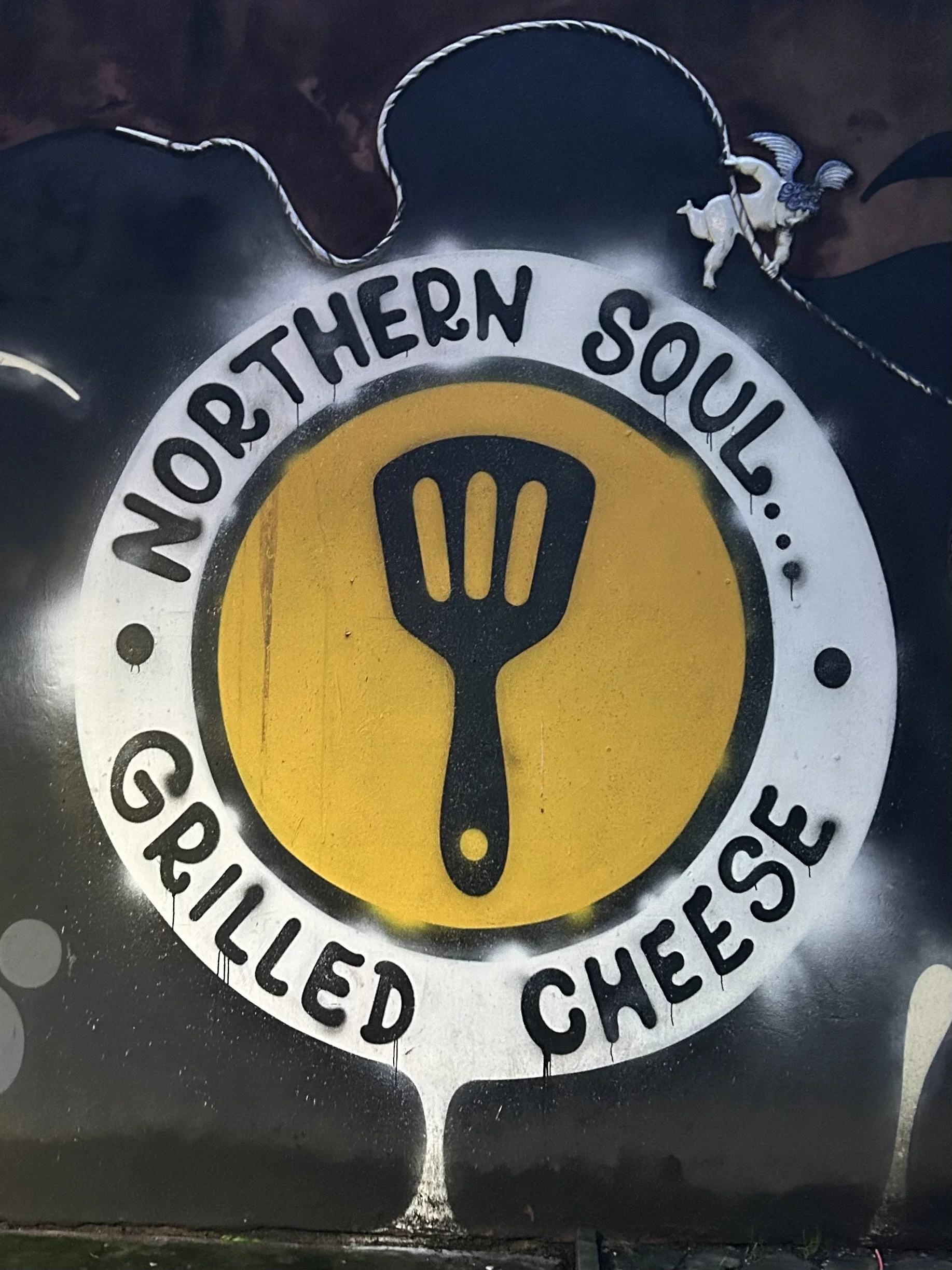 Graffiti art depicting the words 'Northern Soul' and 'Grilled Cheese' with a yellow circle containing a black spatula icon, and a small cartoon character of a pig holding a skewer in the top right corner.