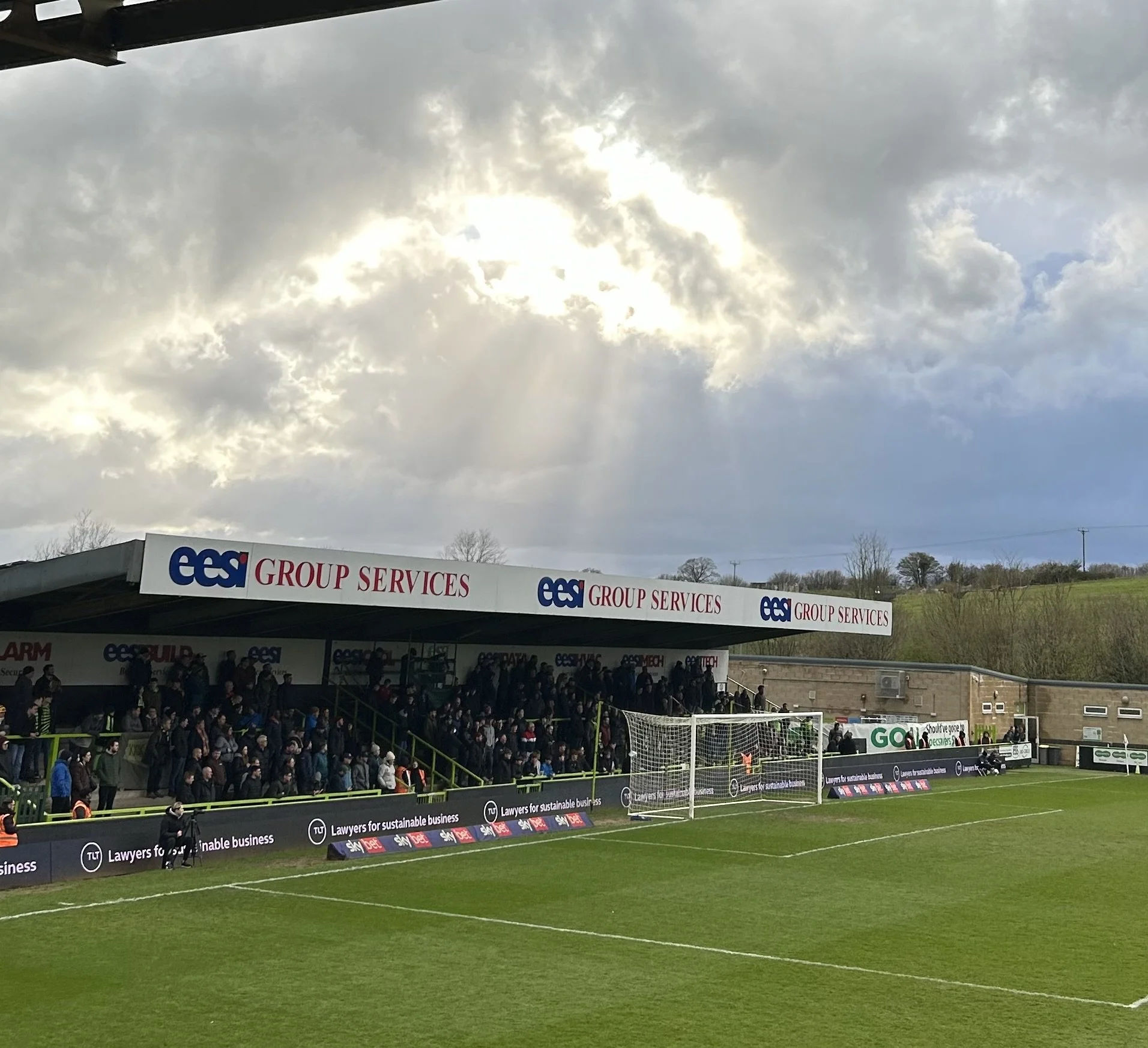 Spectators watching a soccer game from a covered stand at a stadium, with a cloudy sky and sun rays breaking through the clouds.