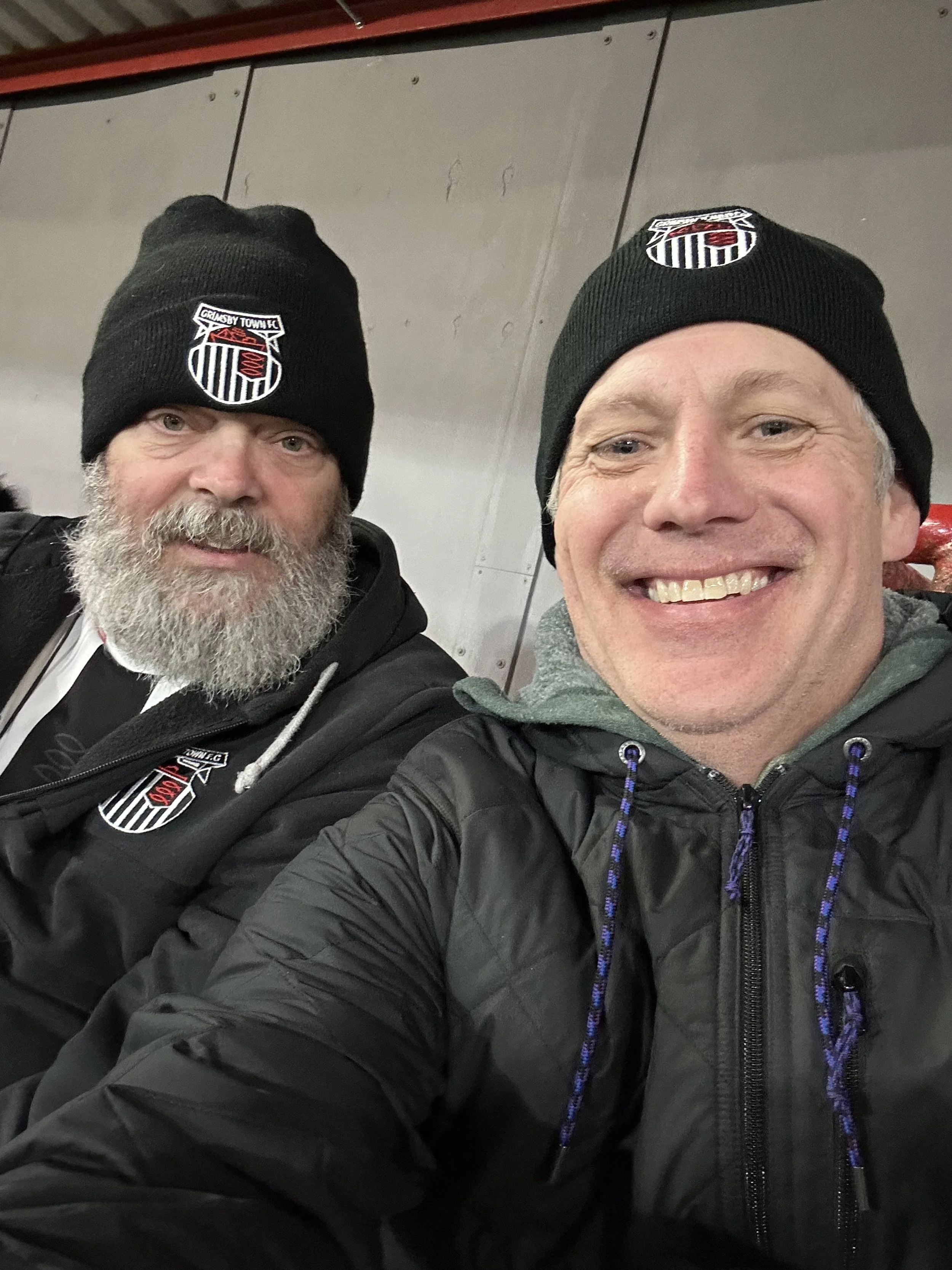 Two men wearing black beanie hats with a logo, smiling for a selfie in a stadium or arena with a gray metal wall in the background.