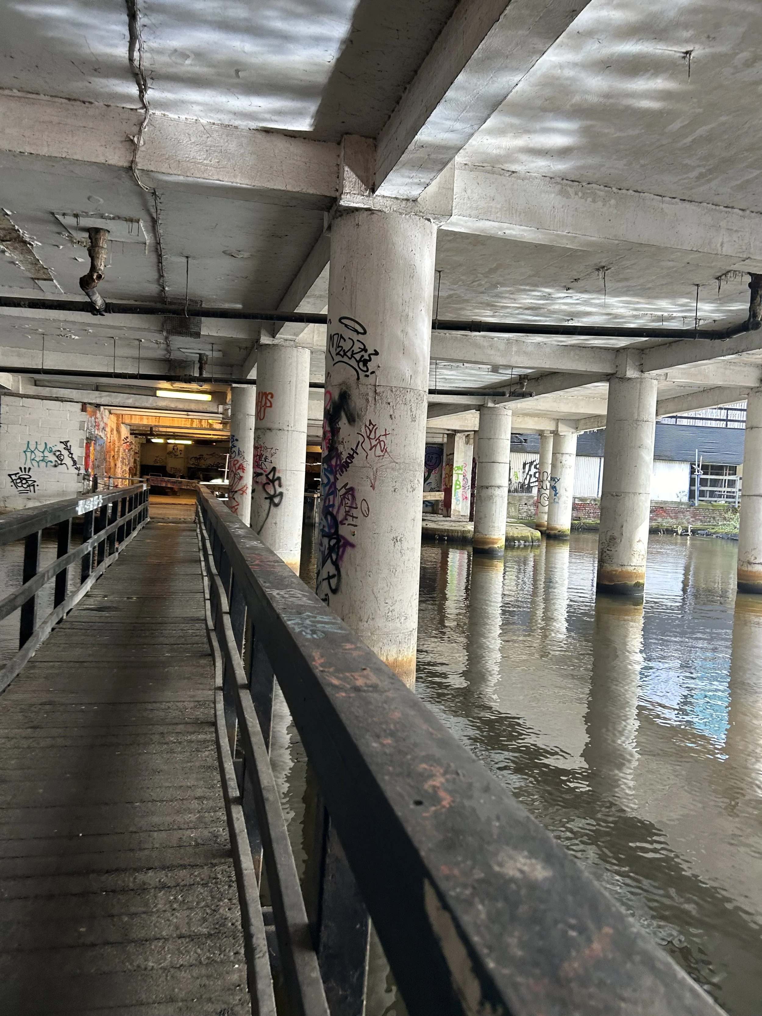 Under a concrete bridge over water, a walkway with a black metal railing, graffiti on the columns, and an opening in the bridge leading to a lit area in the distance.