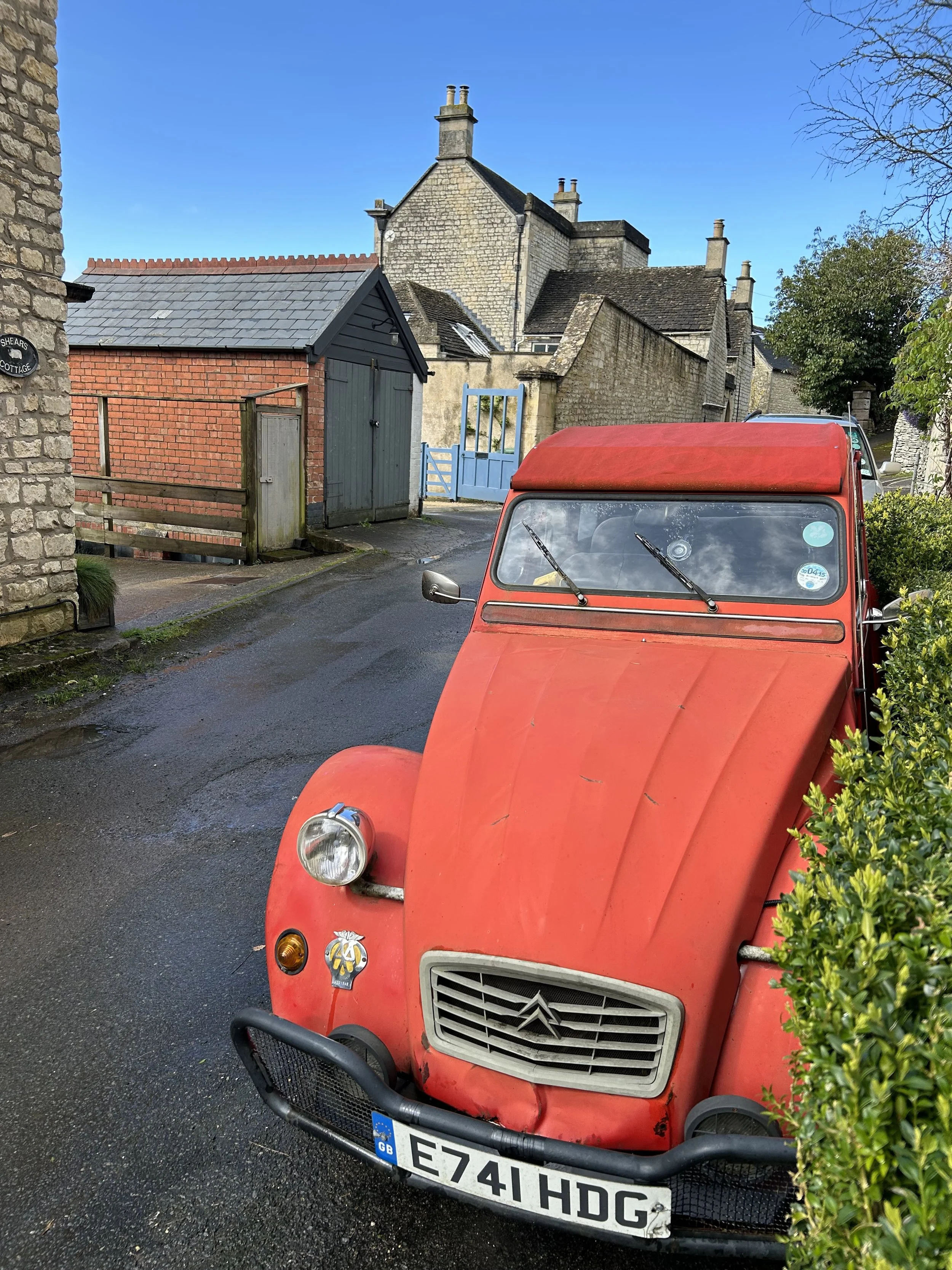 A red vintage Citroën 2CV parked on a narrow street with stone buildings and a blue gate in the background.