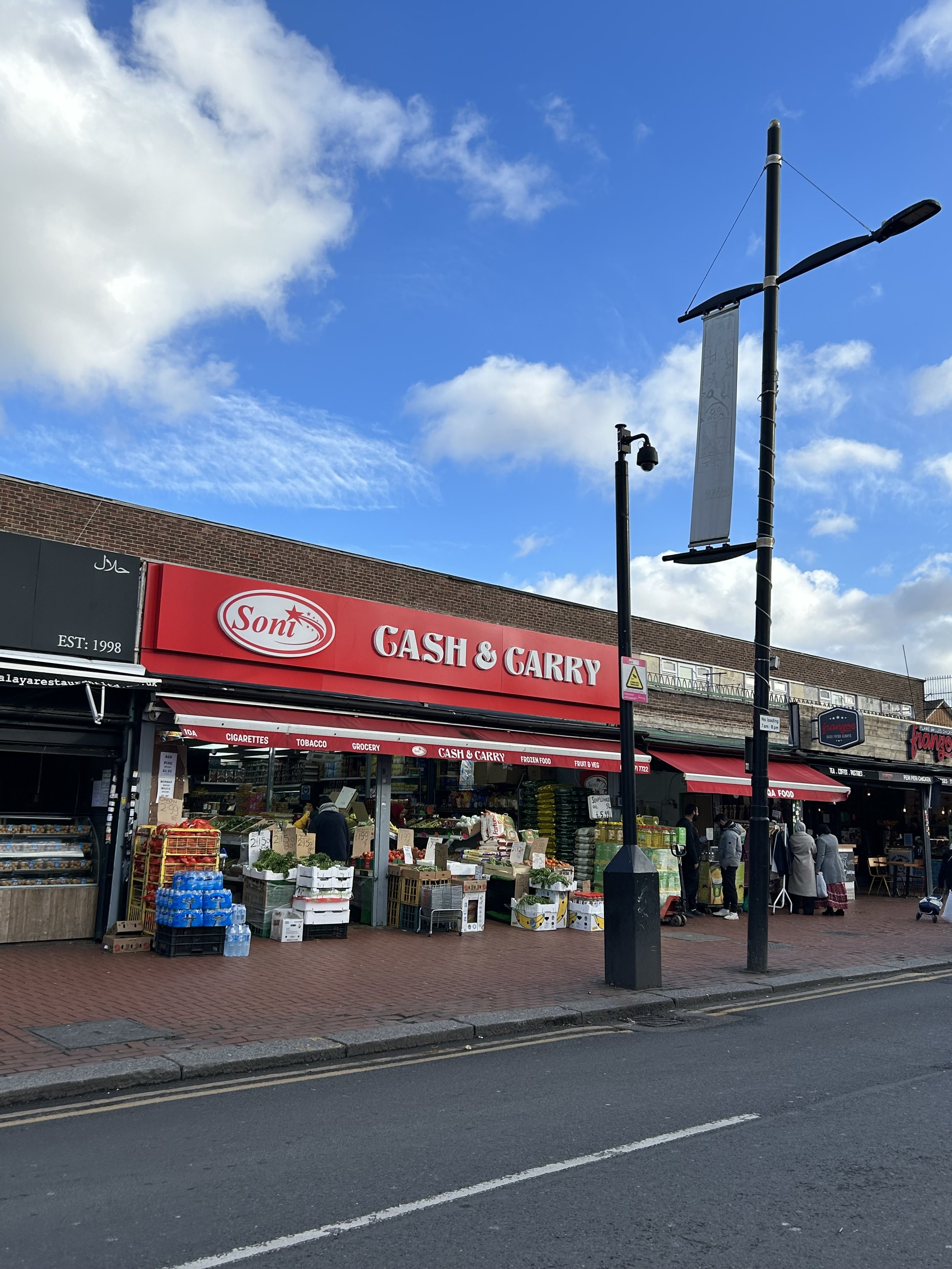 Street view of a grocery store named 'Sont' with a red sign, selling groceries, fruits, and vegetables. Several people are shopping outside under a partly cloudy sky.