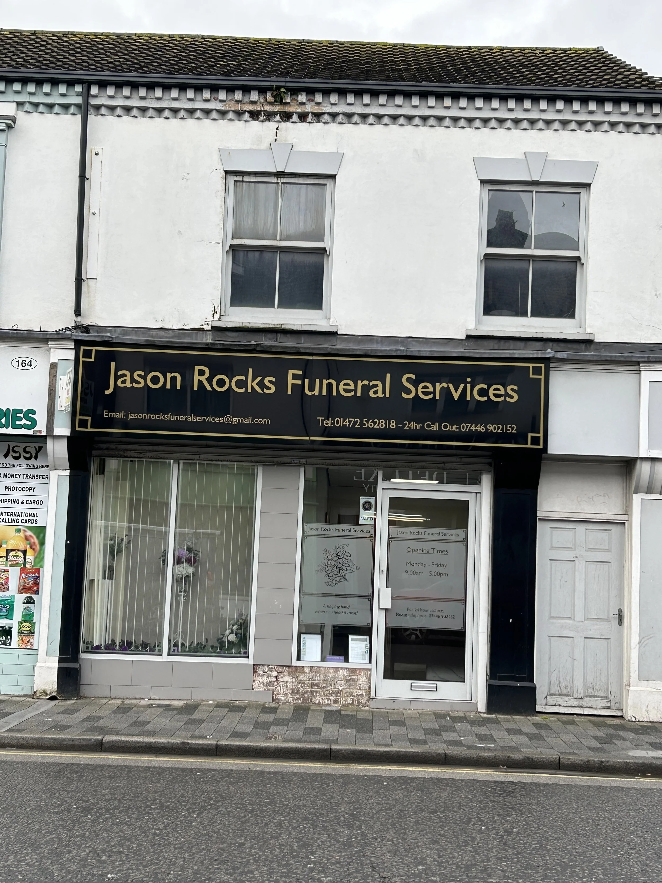  storefront with a sign reading 'Jason Rocks Funeral Services'; the building has two windows on the upper floor and the ground floor has a door and window display of flowers, with a small brick area at the base of the door.